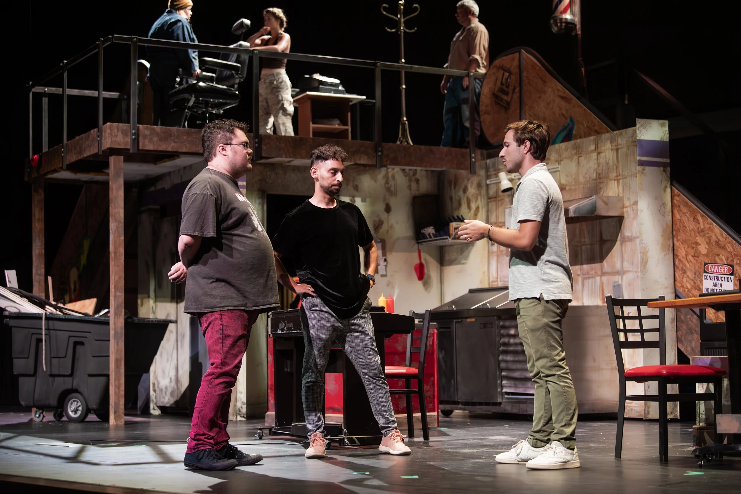 Three men are standing and talking in a theatrical stage play setting, with a set resembling a workshop or kitchen, and two people on a balcony above.