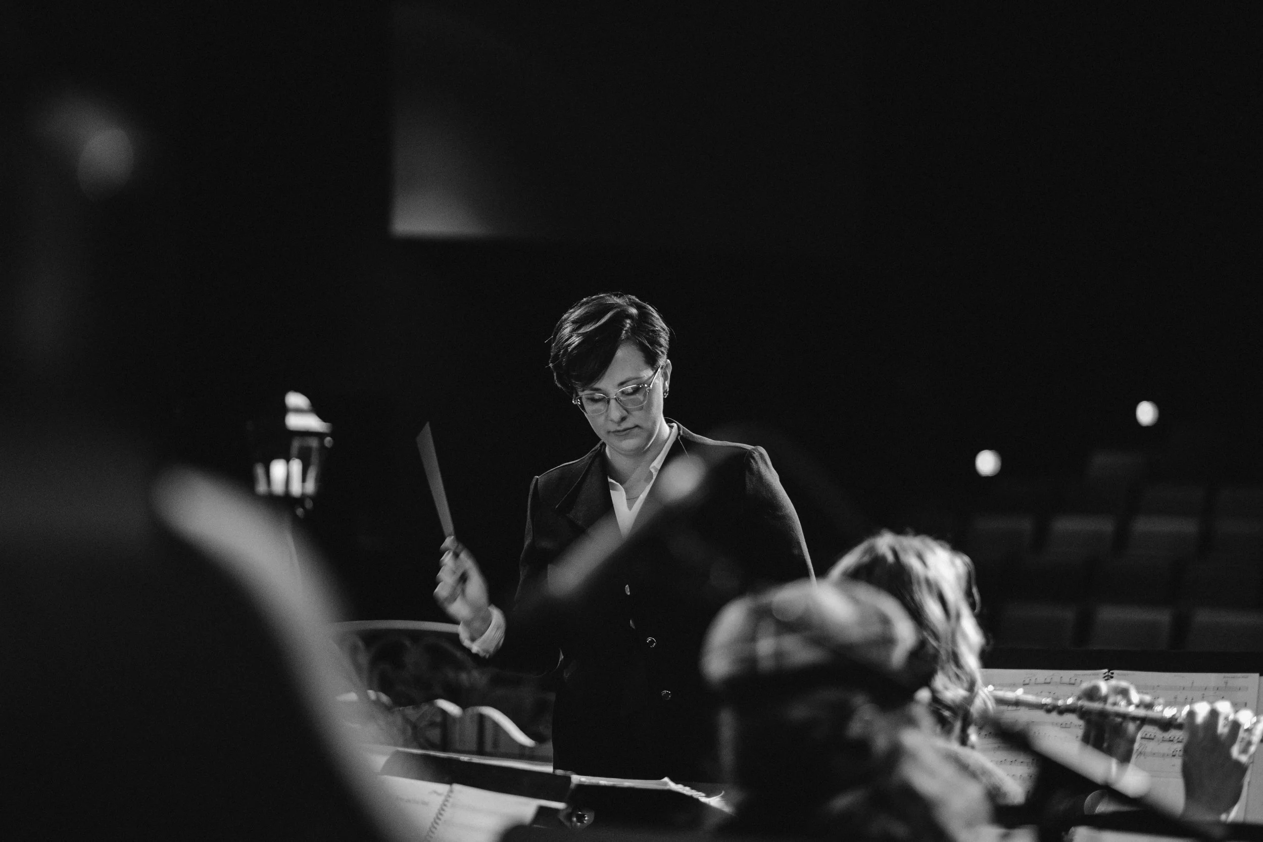 A black-and-white photo of a woman conducting an orchestra with a baton, focusing on her face and the musician in front of her playing a flute.