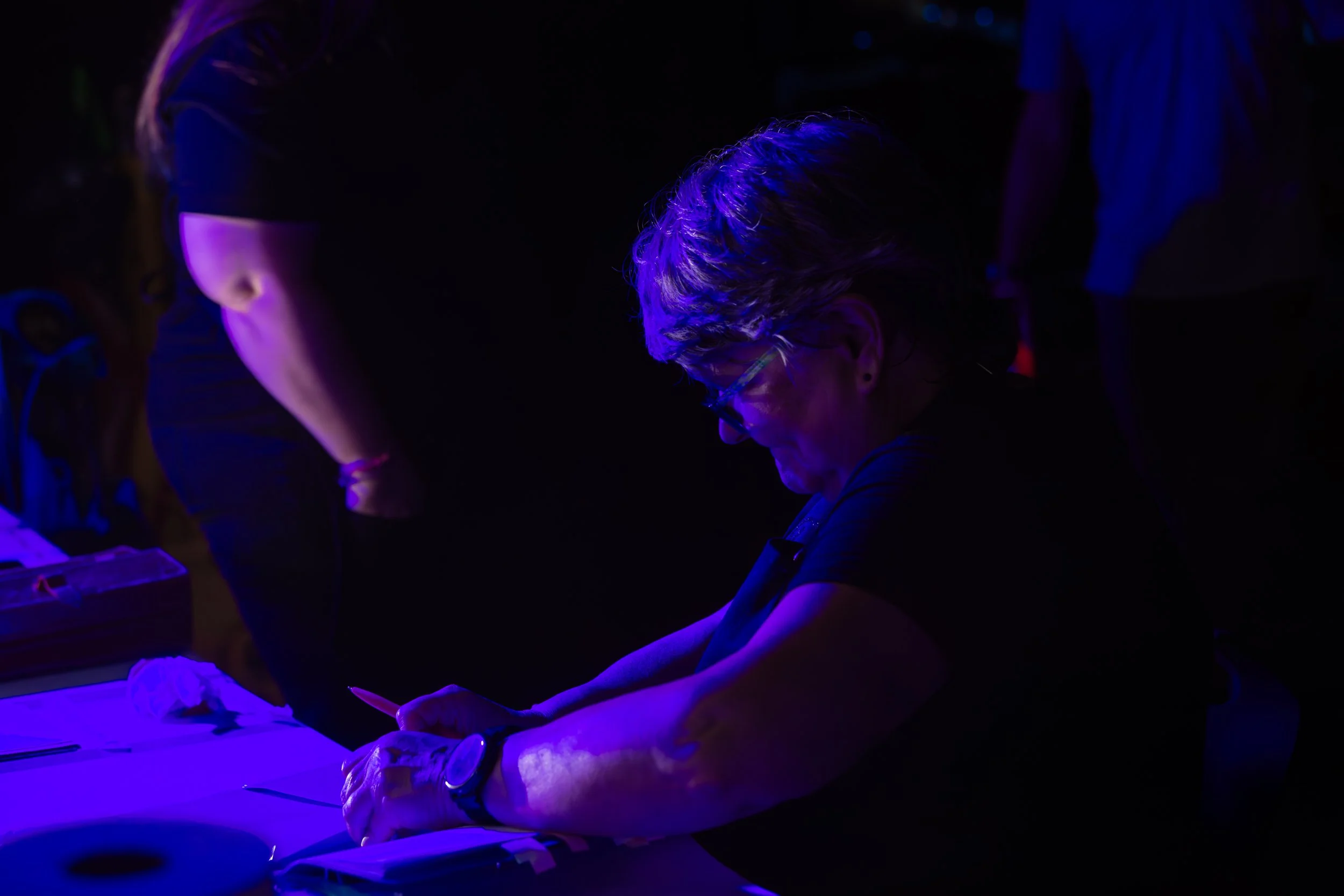 A woman with glasses is writing on a piece of paper at a table in a dark environment illuminated by purple and blue lights.