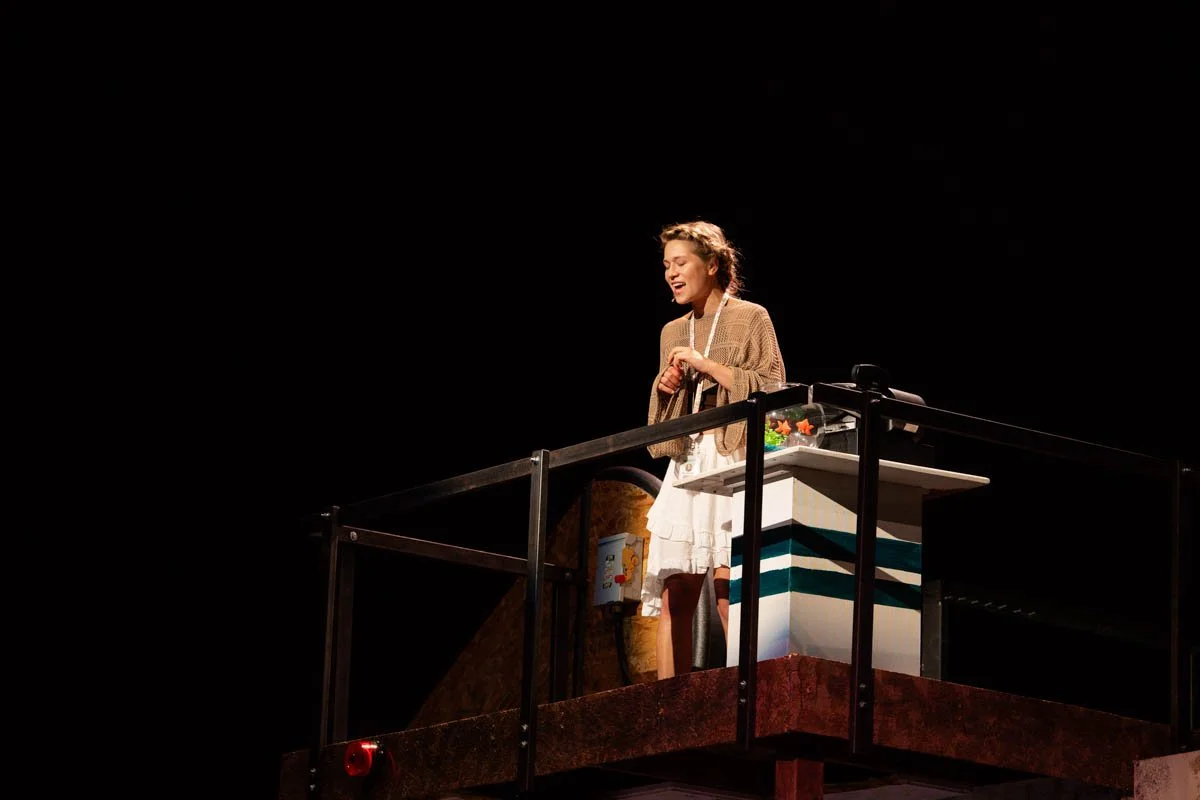 A woman stands on a raised platform with a black railing, singing or speaking, with a plain black background.