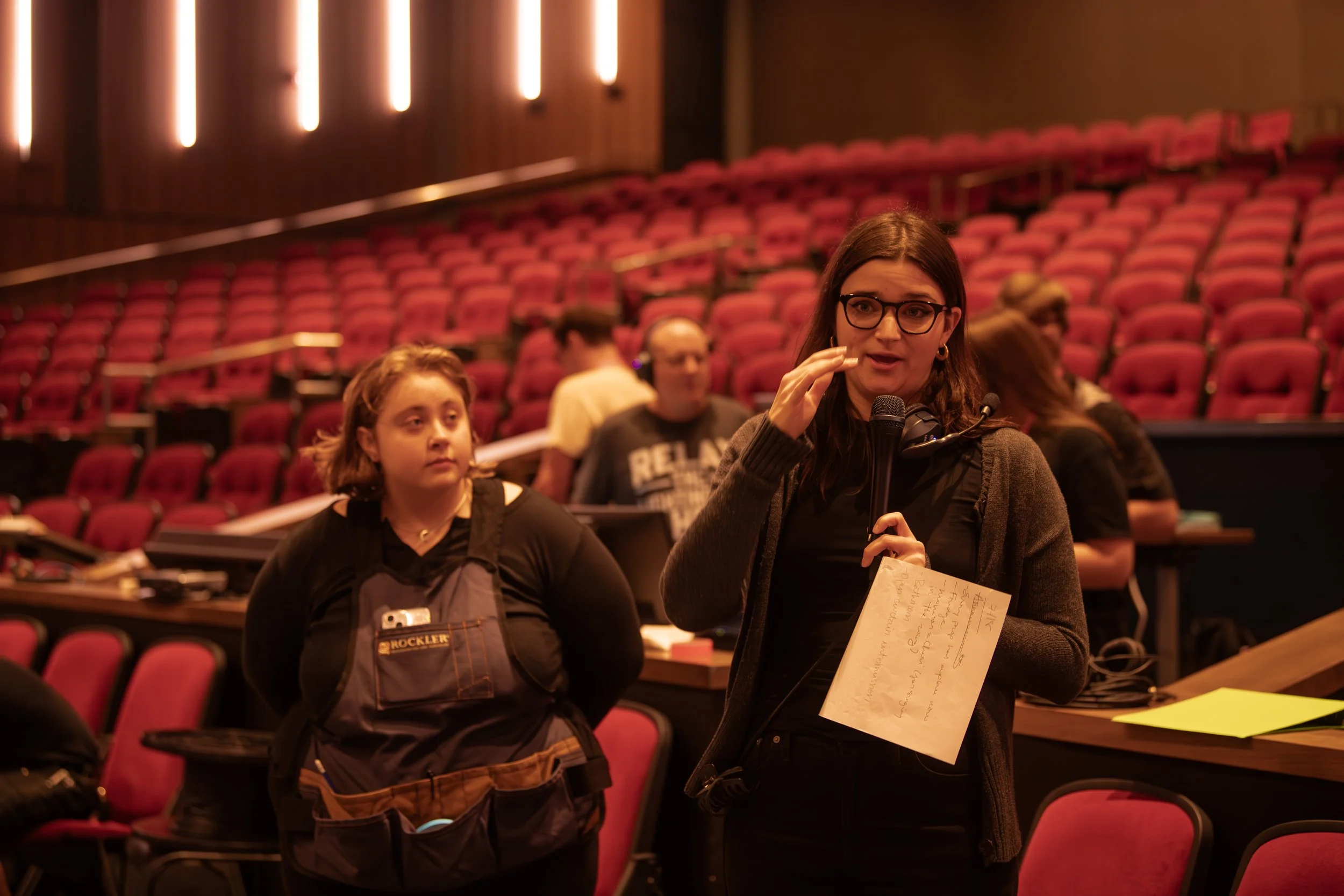 A woman with glasses speaking into a microphone at a conference or meeting in a theater with red seats. Several people are seated in the background.