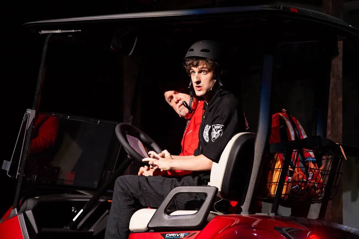 A young man dressed as a security guard or police officer, wearing a black uniform and helmet, sitting in a red golf cart with an older man sitting beside him, both looking surprised or worried, with bags attached to the back of the golf cart.