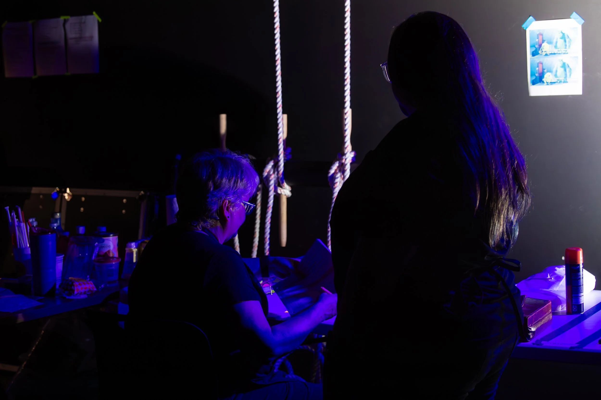 Two women with glasses working at a table illuminated by blue and purple lighting, with ropes hanging on the wall and photo prints taped to the wall.