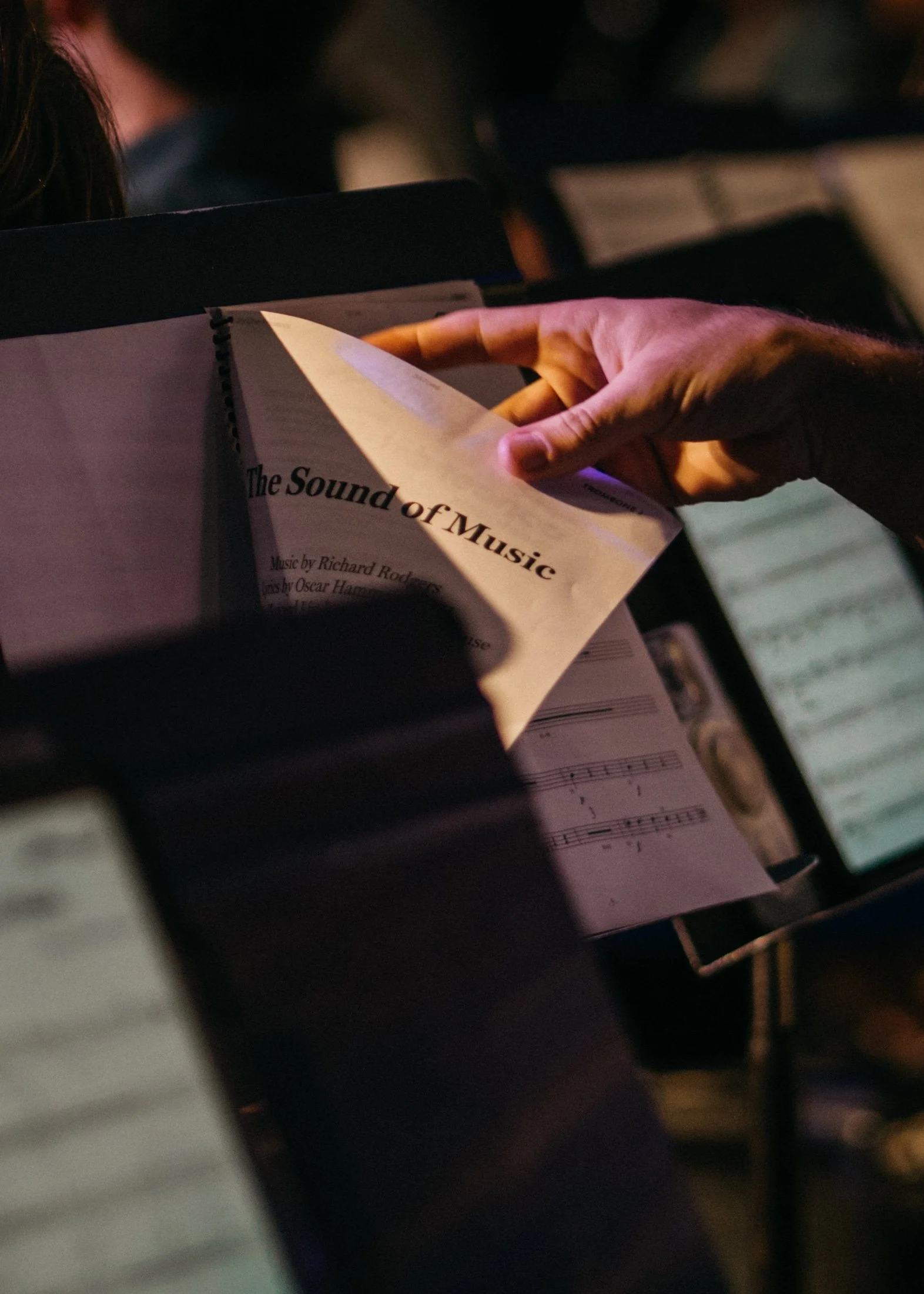 A person flipping through sheet music titled 'The Sound of Music' during a performance or practice, with other sheets and music stands visible in the background.