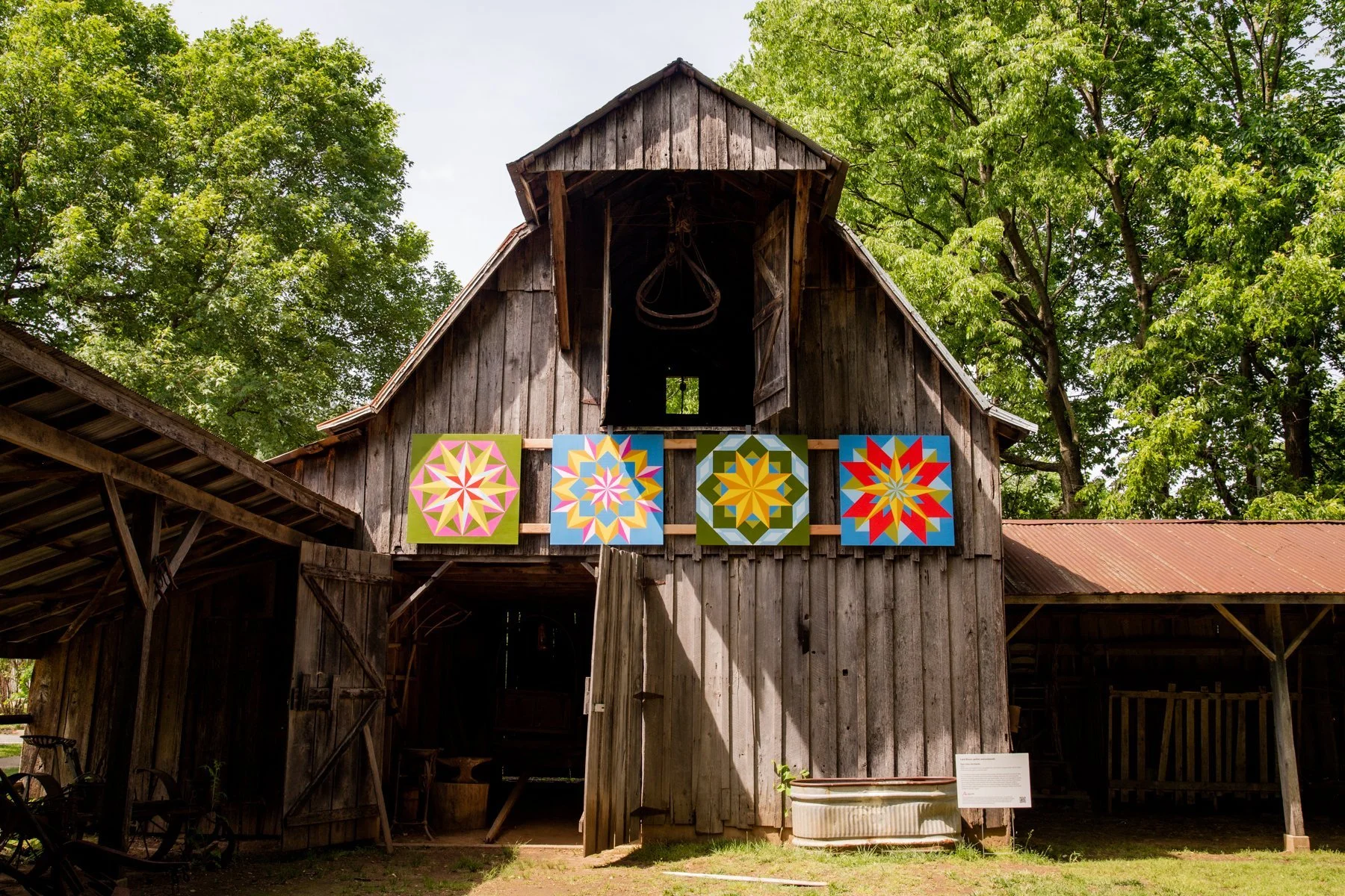 Four colorful geometric quilt patterns are displayed on the front of a rustic wooden barn with a partially open door, surrounded by green trees.