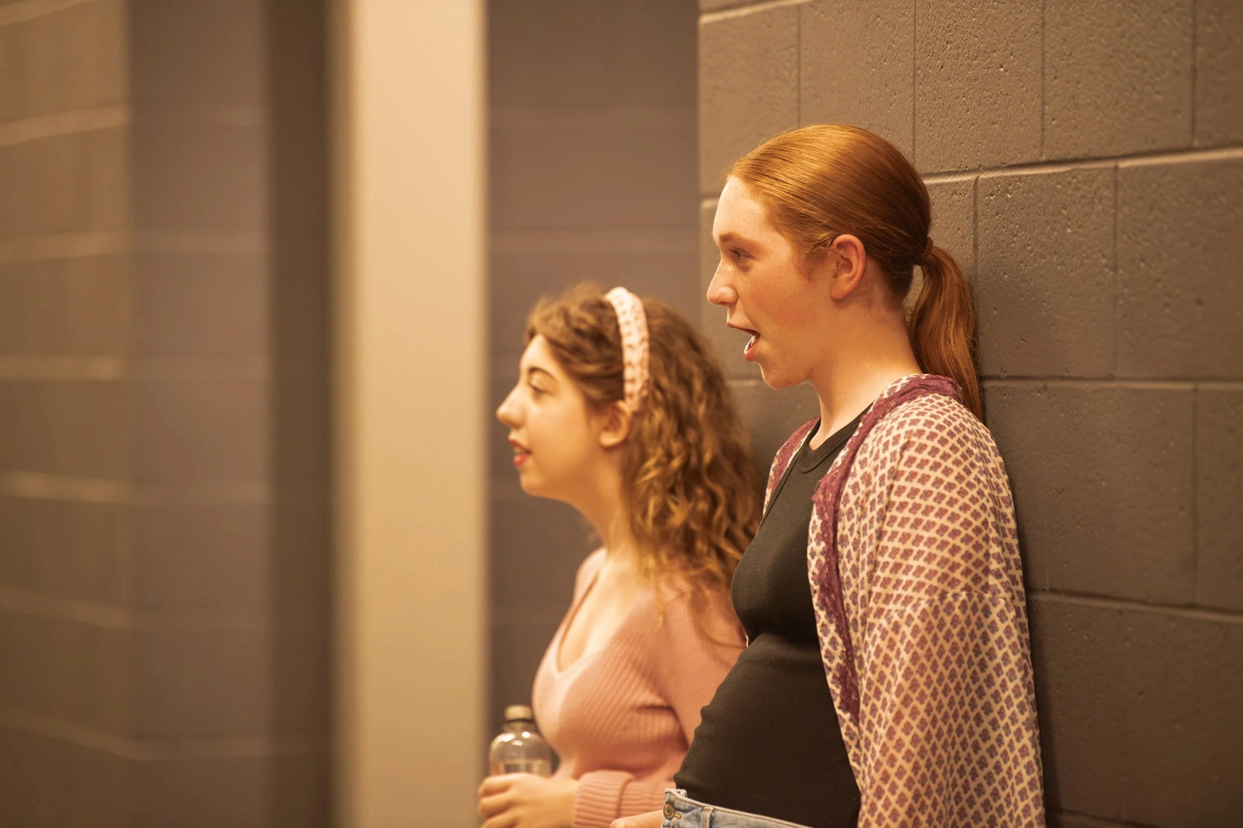 Two young women standing against a gray wall, one with red hair and the other with brown, both engaged in conversation.