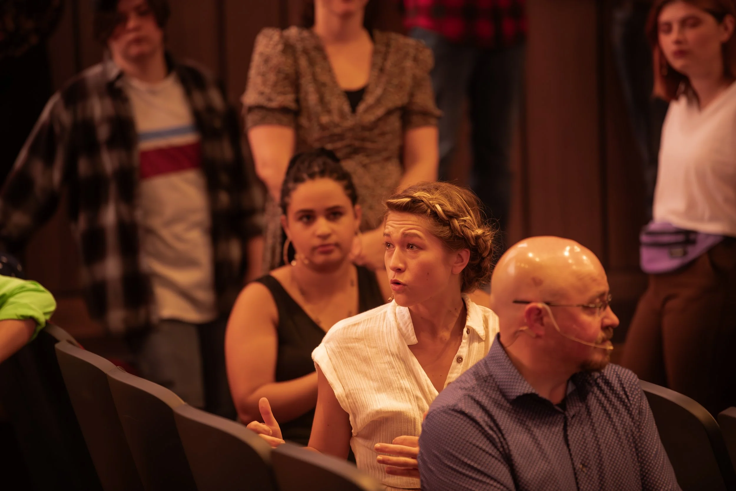 A group of people seated and standing, engaged in a discussion or listening during a meeting or event in a dimly lit room. One woman with short blonde hair and a white shirt is speaking, while others look on attentively.