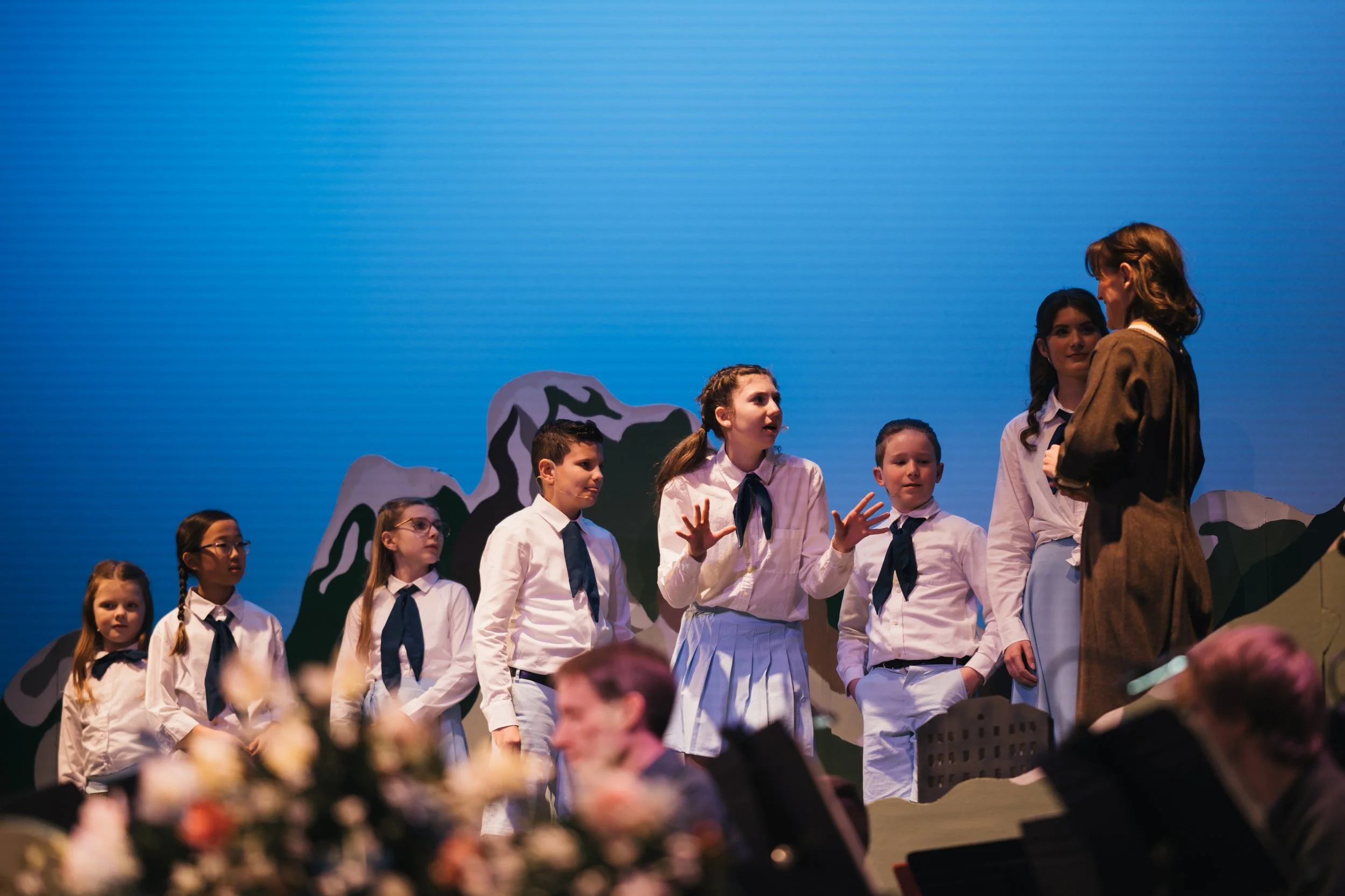 Children performing on stage in school uniforms with blue ties, engaging in a dialogue with a woman dressed in a brown costume, against a backdrop of painted mountainous scenery.