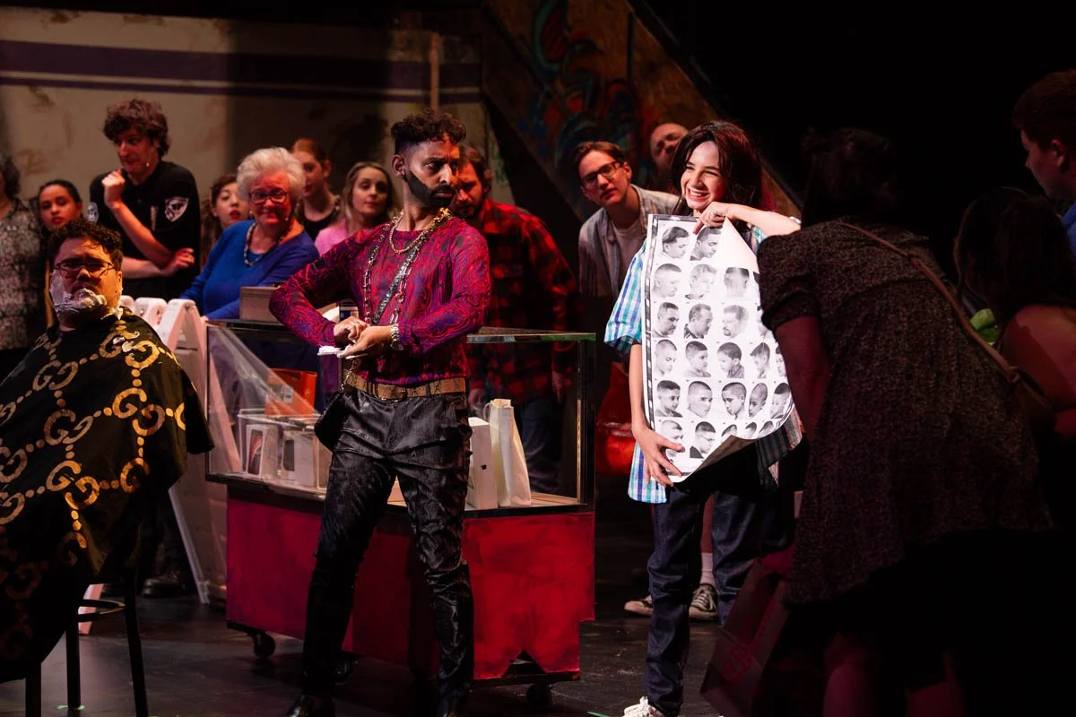 A lively scene with several people, including a woman smiling and holding a large book with black-and-white photos of a person's different hairstyles. A man in colorful clothing stands near a glass display case. The background features a group of onl
