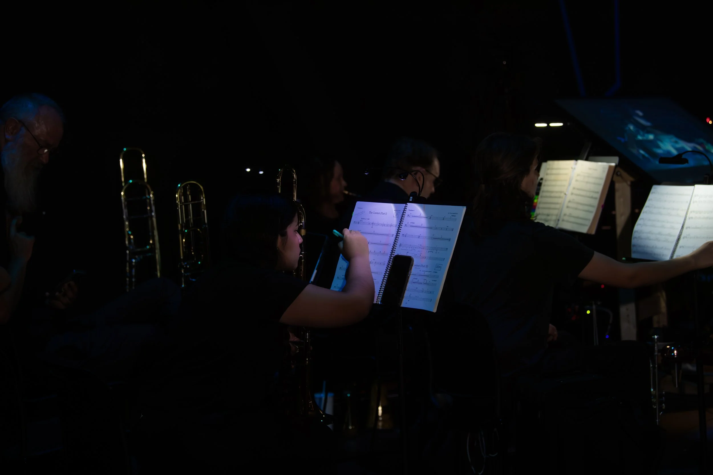Orchestra members, including a young girl, seated with music stands and sheet music in a dark concert hall.