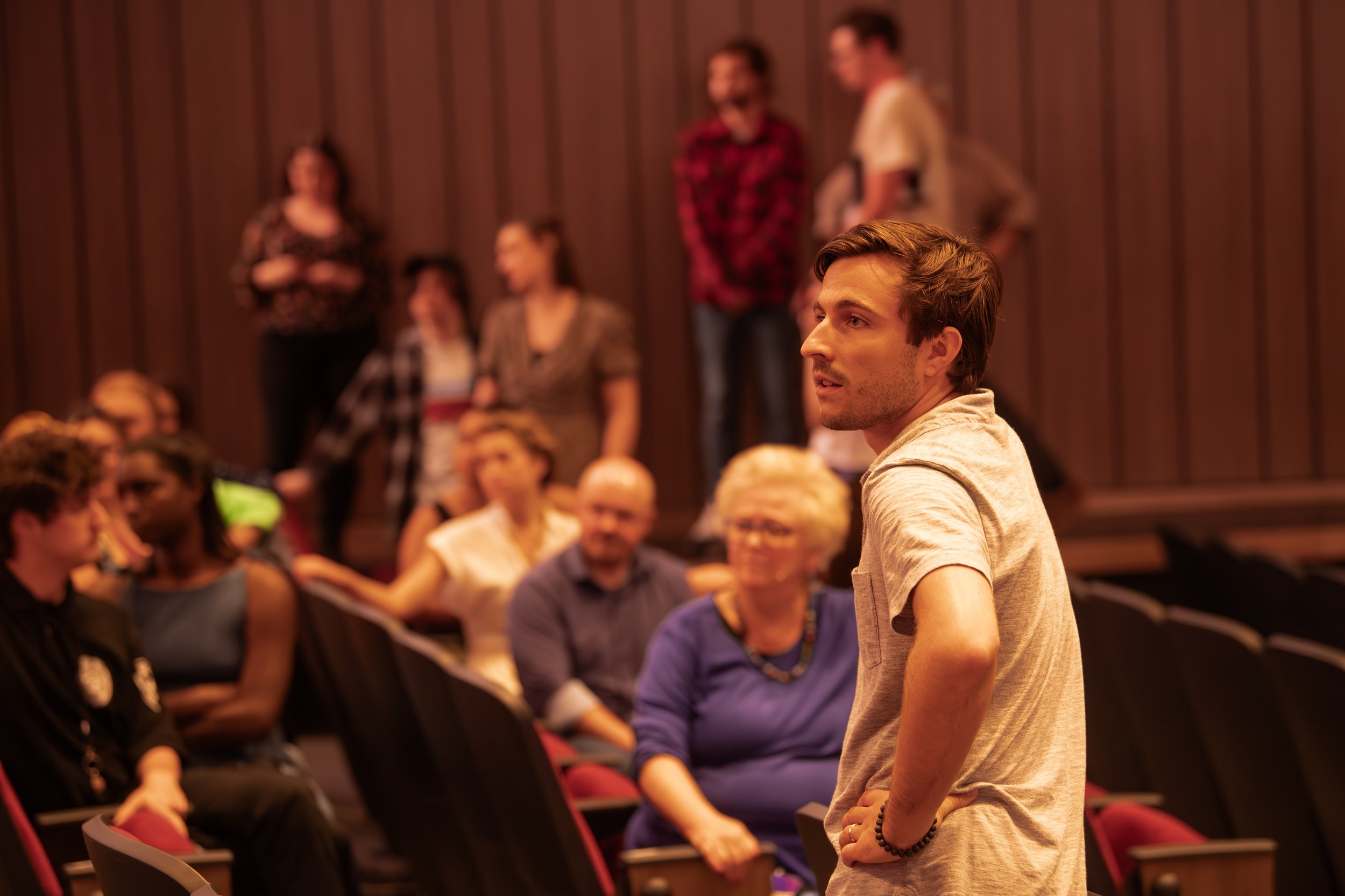 A young man in a beige t-shirt stands with his hand on his hip, looking to the side, while a group of people sit and stand around him in a room with wood-paneled walls.