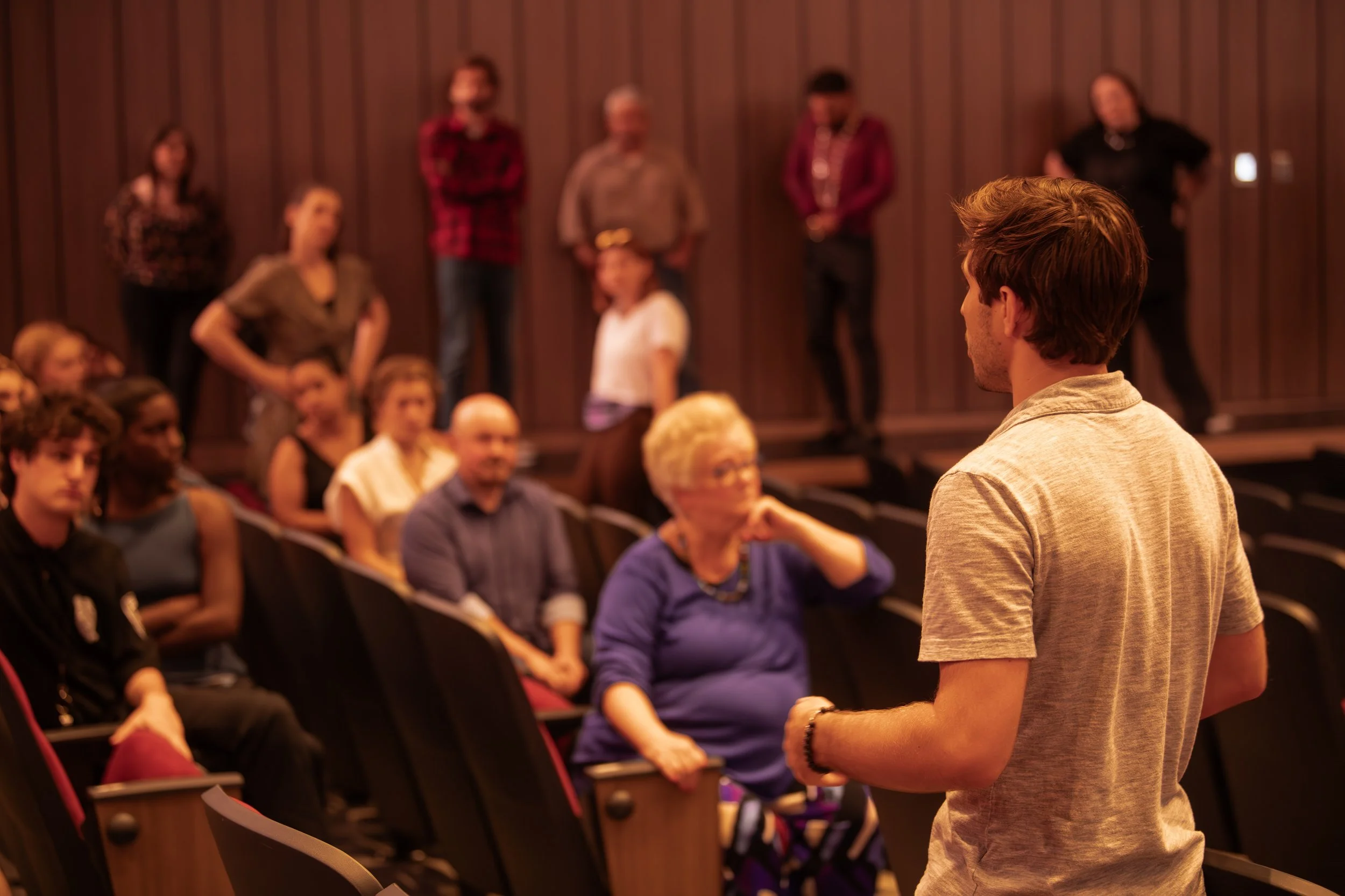 A man with brown hair wearing a gray shirt is speaking in front of an audience seated in a theater or auditorium with brown wood paneling.