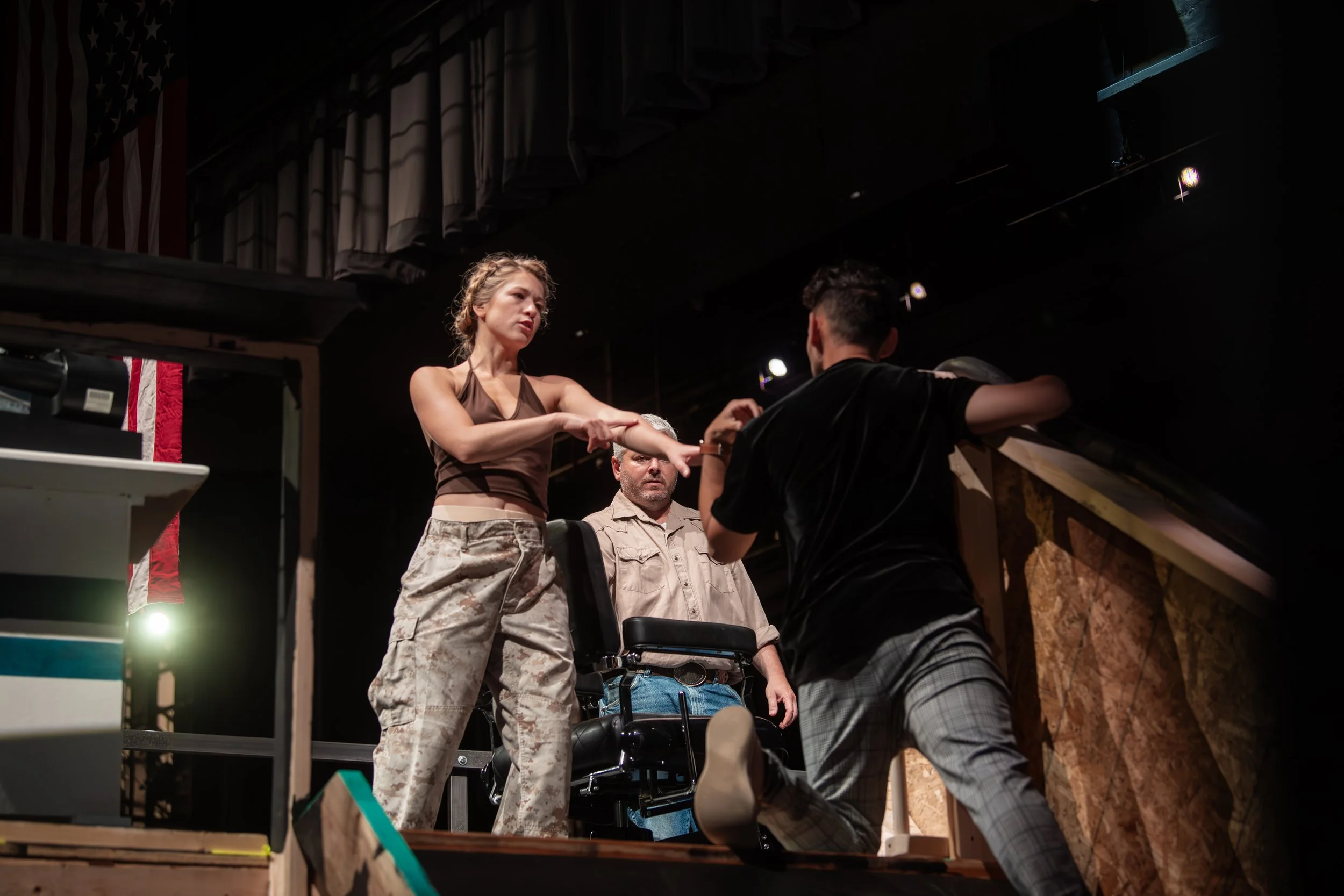 A scene from a theatrical performance shows a woman with her arms extended, appearing to act or argue with a man kneeling in front of her. A third person, an older man, stands behind them observing. The setting appears to be a stage with dim lighting