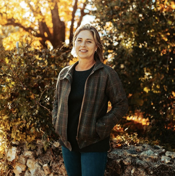 A woman standing outdoors in autumn, smiling, wearing a plaid jacket, black shirt, and blue jeans, with trees and fall foliage in the background.