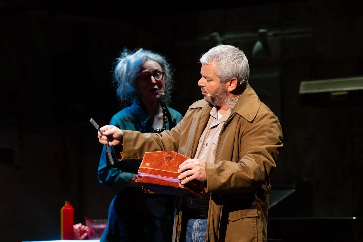 A man with gray hair wearing a brown coat and a woman with curly hair and glasses wearing a blue jacket and necklace on stage, with the man holding a brown case and a pen.