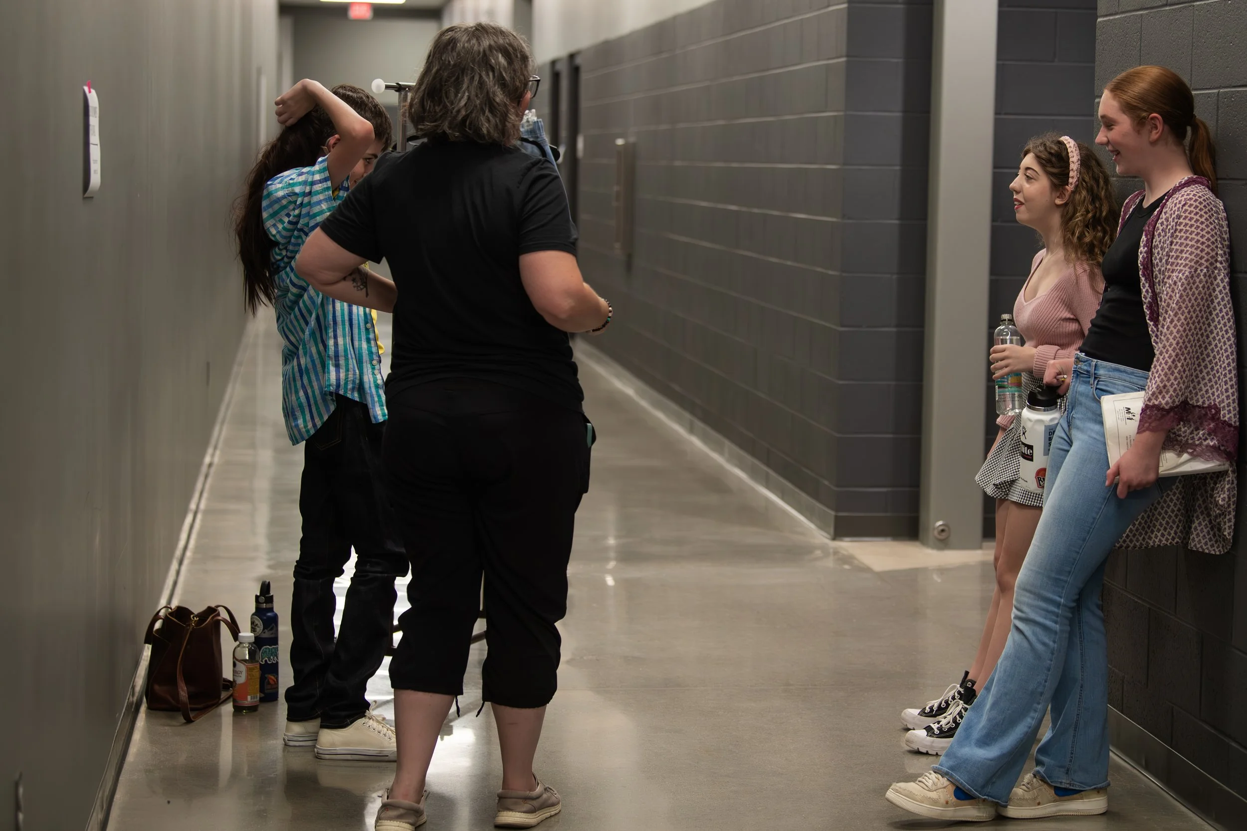 A group of four girls and one woman are standing in a hallway, talking to each other. Two girls are leaning against the wall on the right, one girl is in the middle with her back to the camera, and one girl is on the left, with a woman, all engaged i
