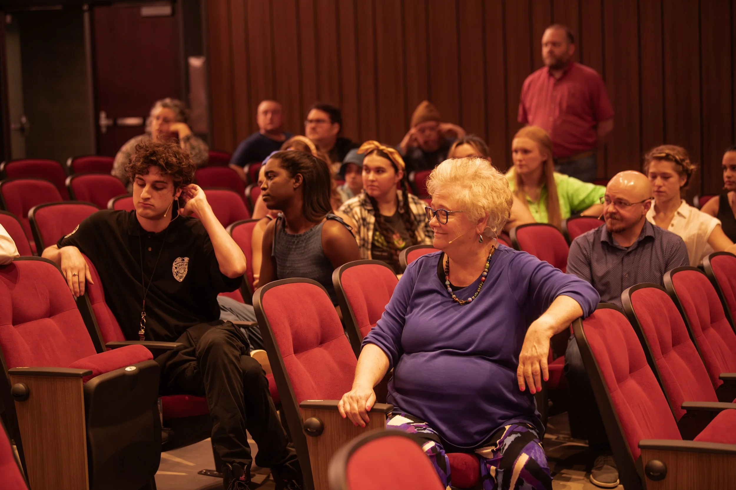 People seated in red theater seats during an indoor event, with some engaging and others waiting.