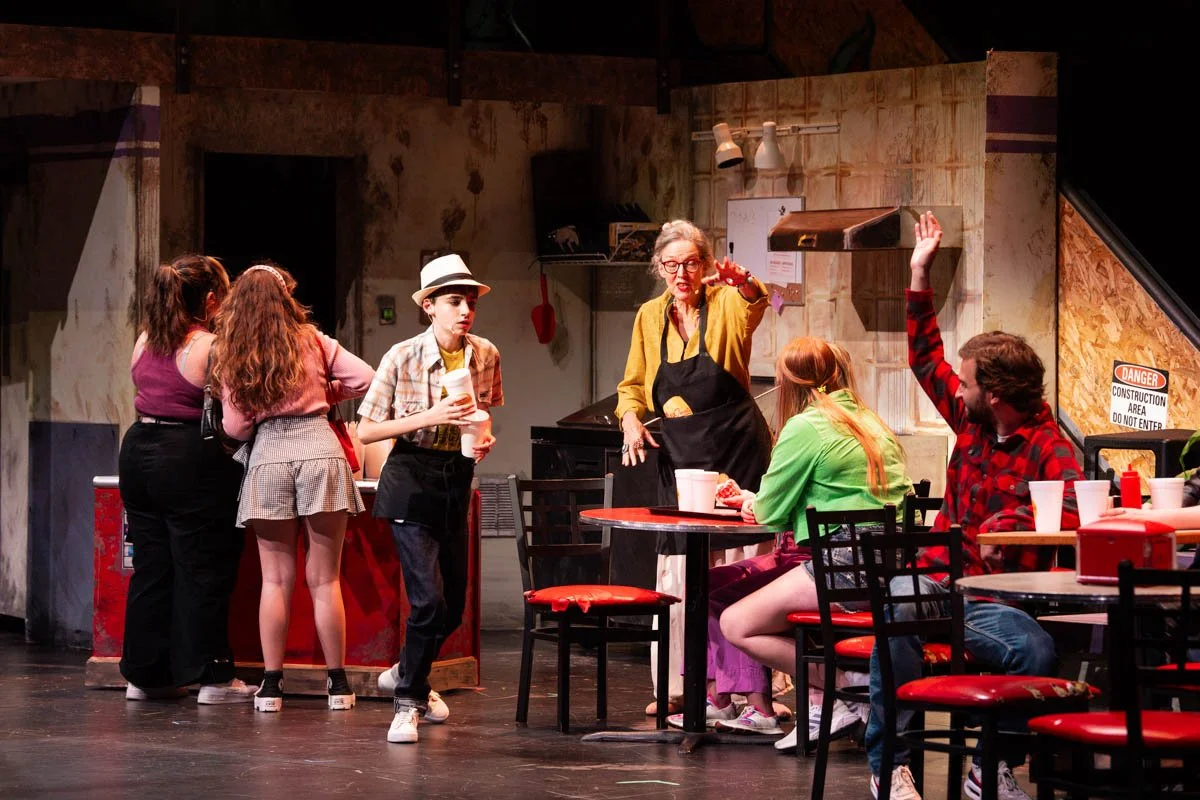 Actors performing in a play set in a casual restaurant or cafe, with some actors seated at tables and others standing, engaging in conversation or acting out a scene.