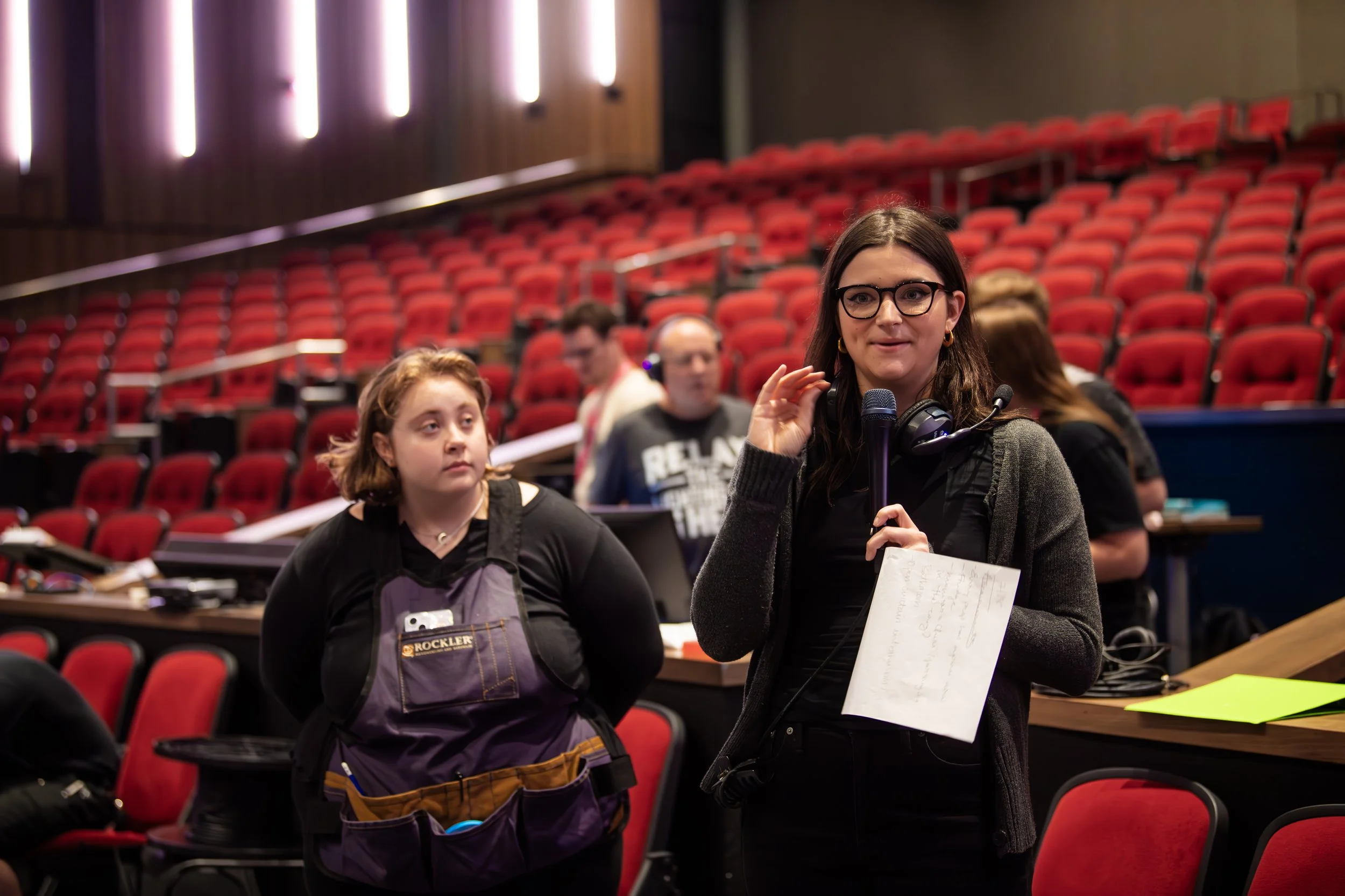 A young woman with glasses holding a microphone and notes speaking on set with people in the background in an auditorium with red seats.