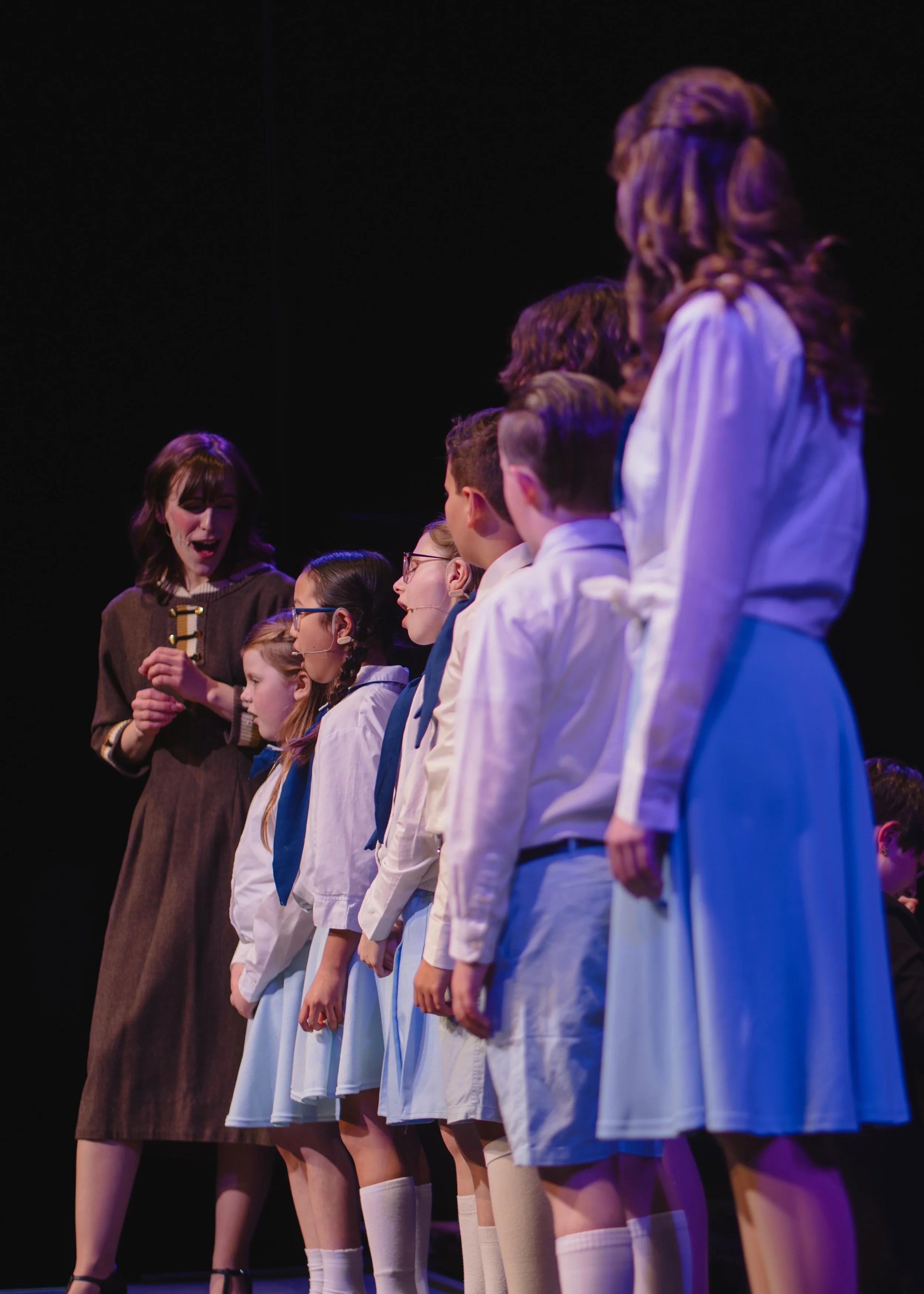 A group of children in school uniforms standing in a line on stage, with a woman speaking to them, possibly during a performance or rehearsal.