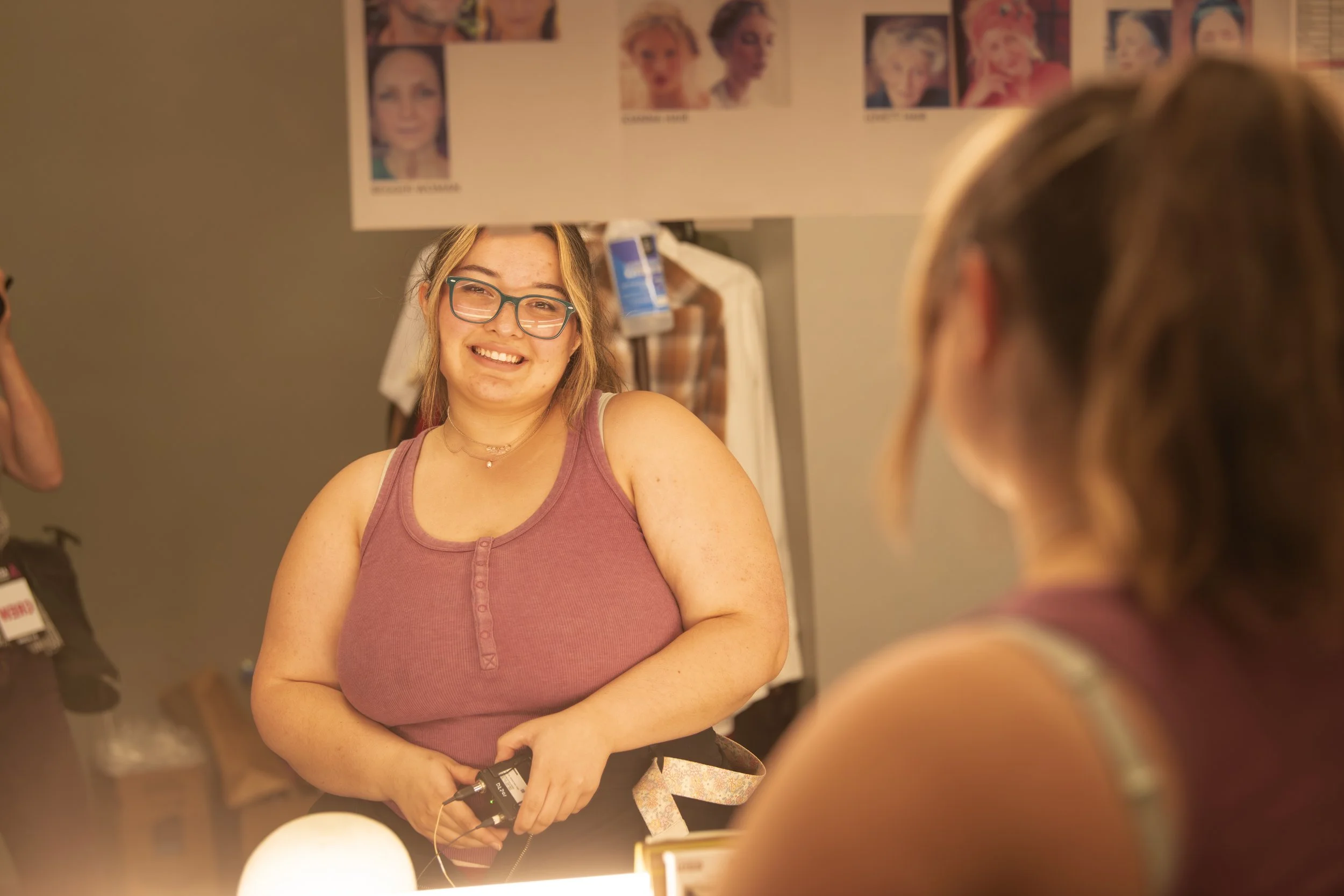 A young woman with glasses smiling while looking at her reflection in a mirror, wearing a sleeveless purple top, in a dressing room with photos of different makeup looks above her head.