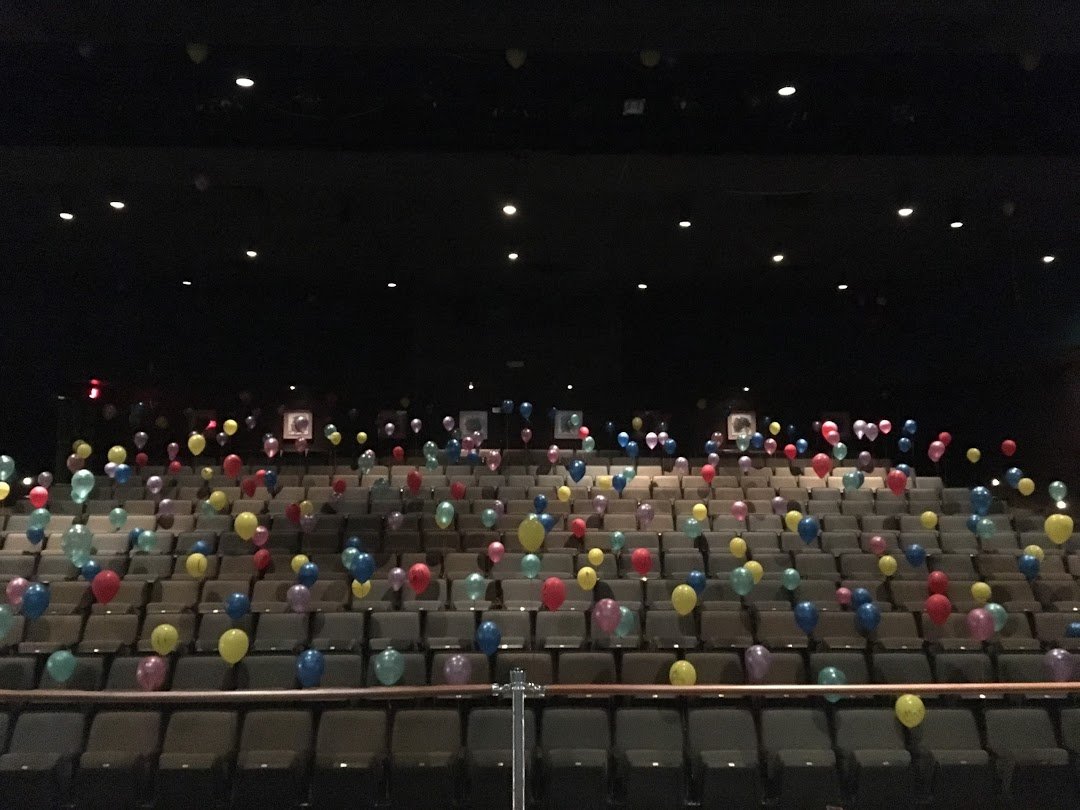 An empty theater with rows of beige seats, decorated with scattered colorful balloons, and framed pictures hanging on the wall in the background.