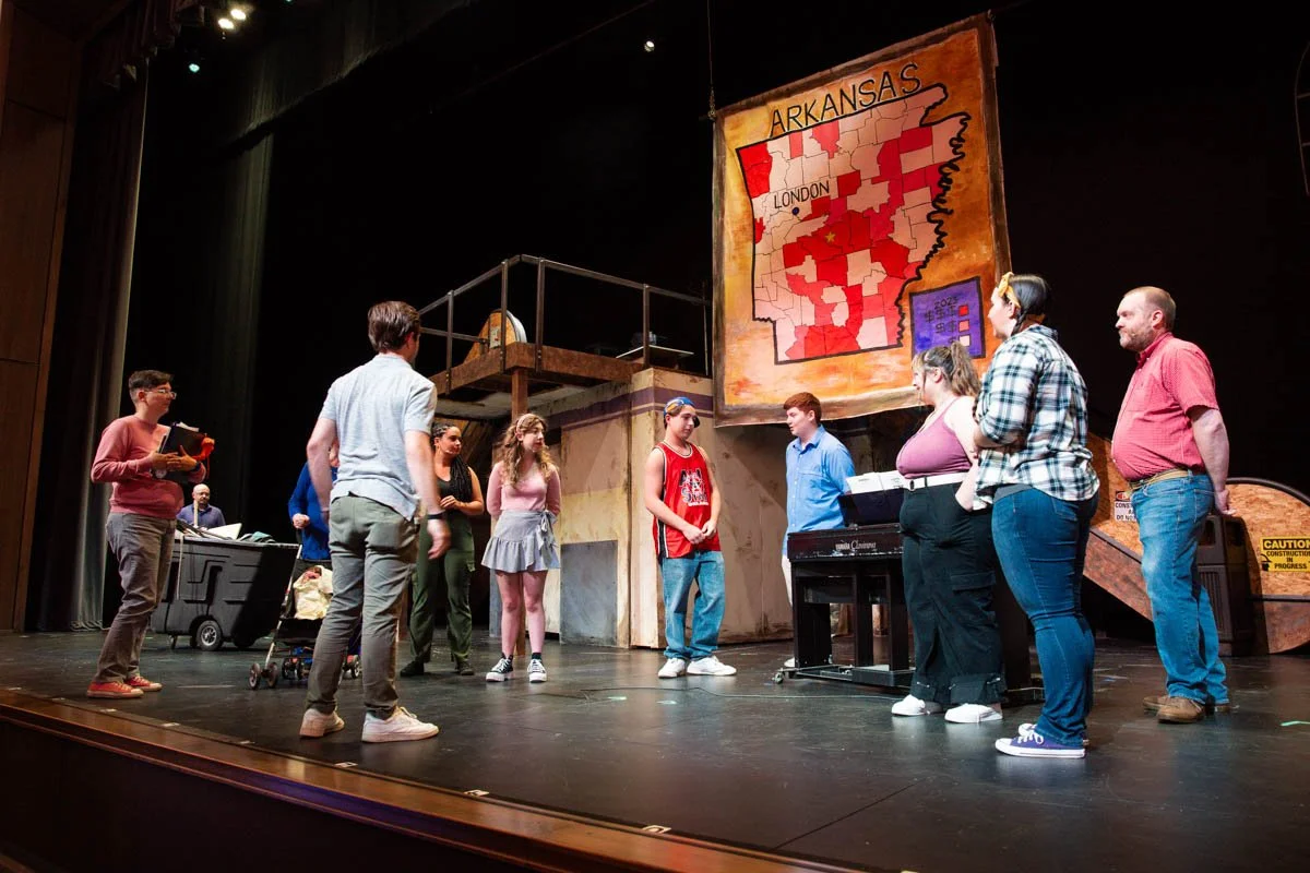 Group of people on a stage rehearsing or performing, with a large map of Arkansas hanging in the background.