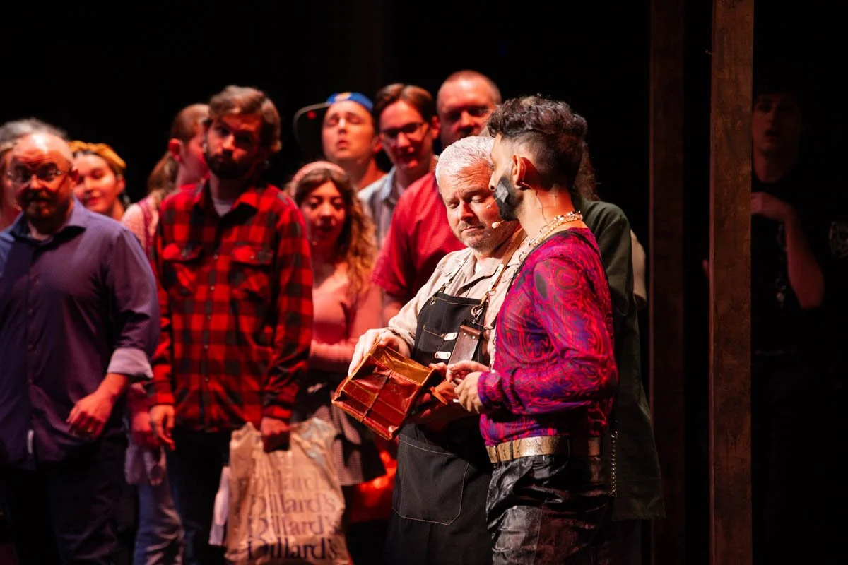 A group of people on stage during a performance, with two actors in the foreground. One actor has gray hair and is wearing an apron, while the other has dark hair styled in a mohawk and is wearing a red patterned jacket. The background features sever