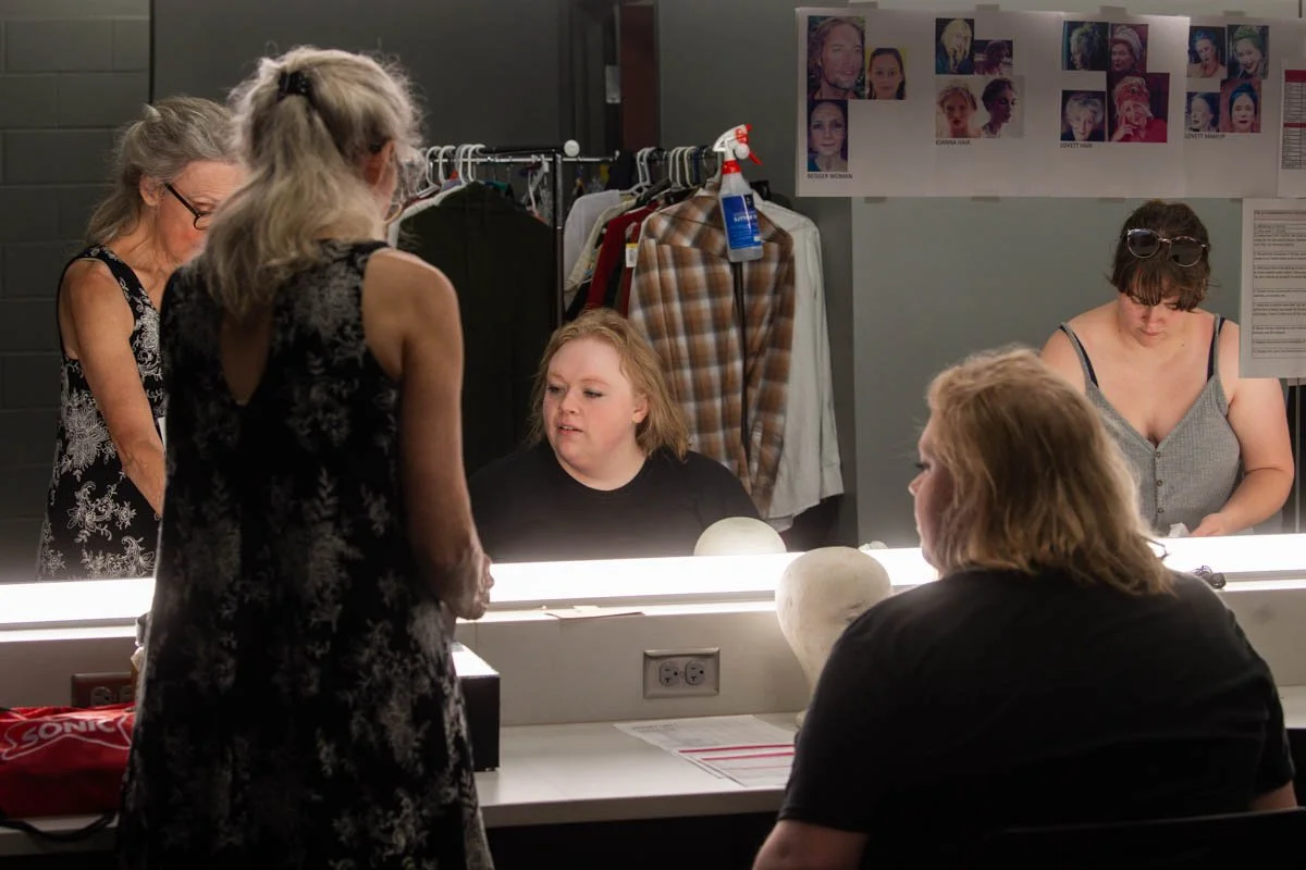 People in a dressing room or makeup room with mirrors, photographs, and clothing racks, preparing for a performance or event.