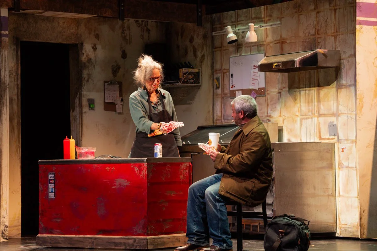 A scene from a play depicting two older characters in a diner or kitchen setting. One woman stands behind a red counter with ketchup and mustard bottles, holding a paper wrapped item. The man sits on a stool holding a wrapped item and a white disposa