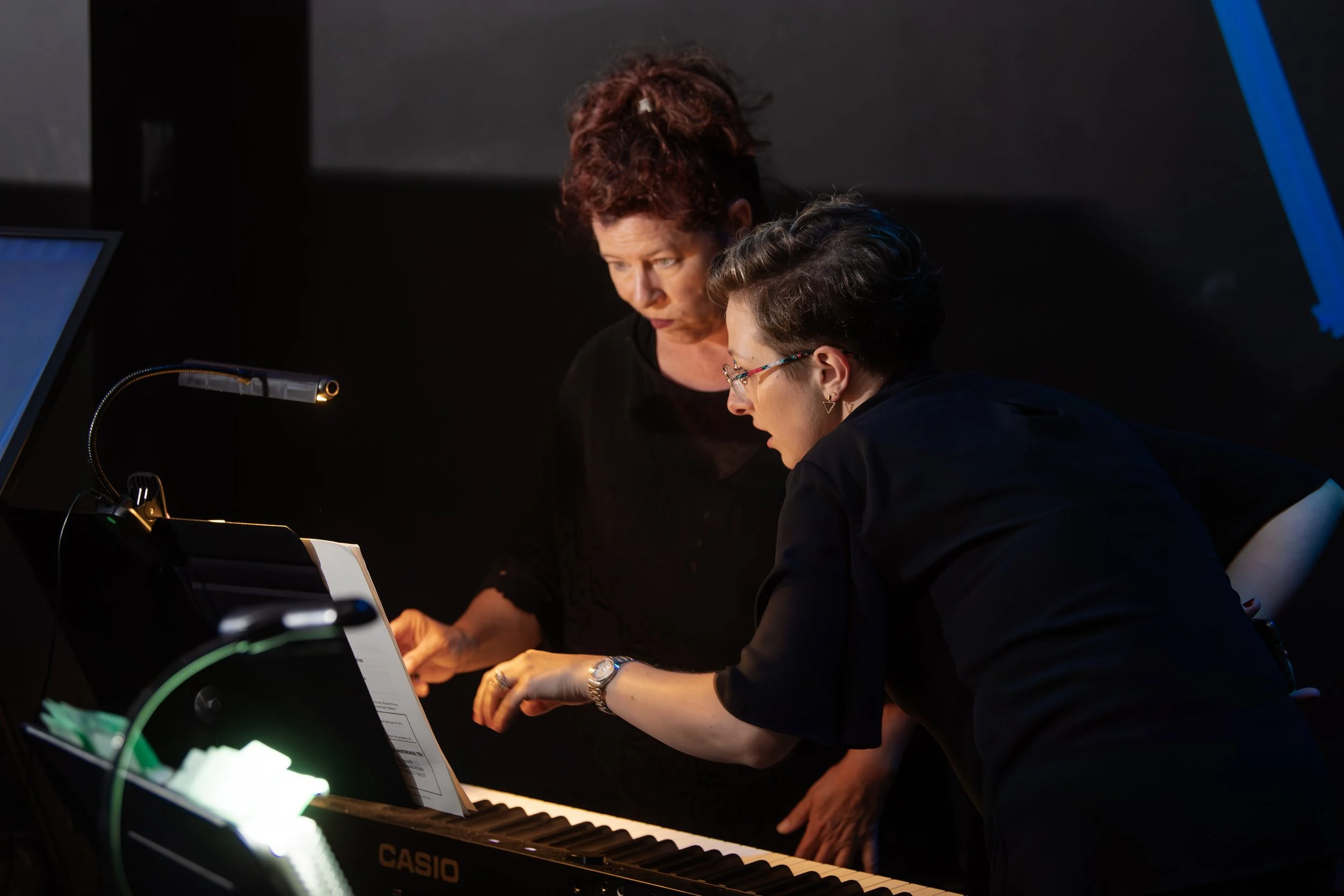 Two women working together on a digital piano, with one woman pointing at sheet music and the other woman looking attentively.