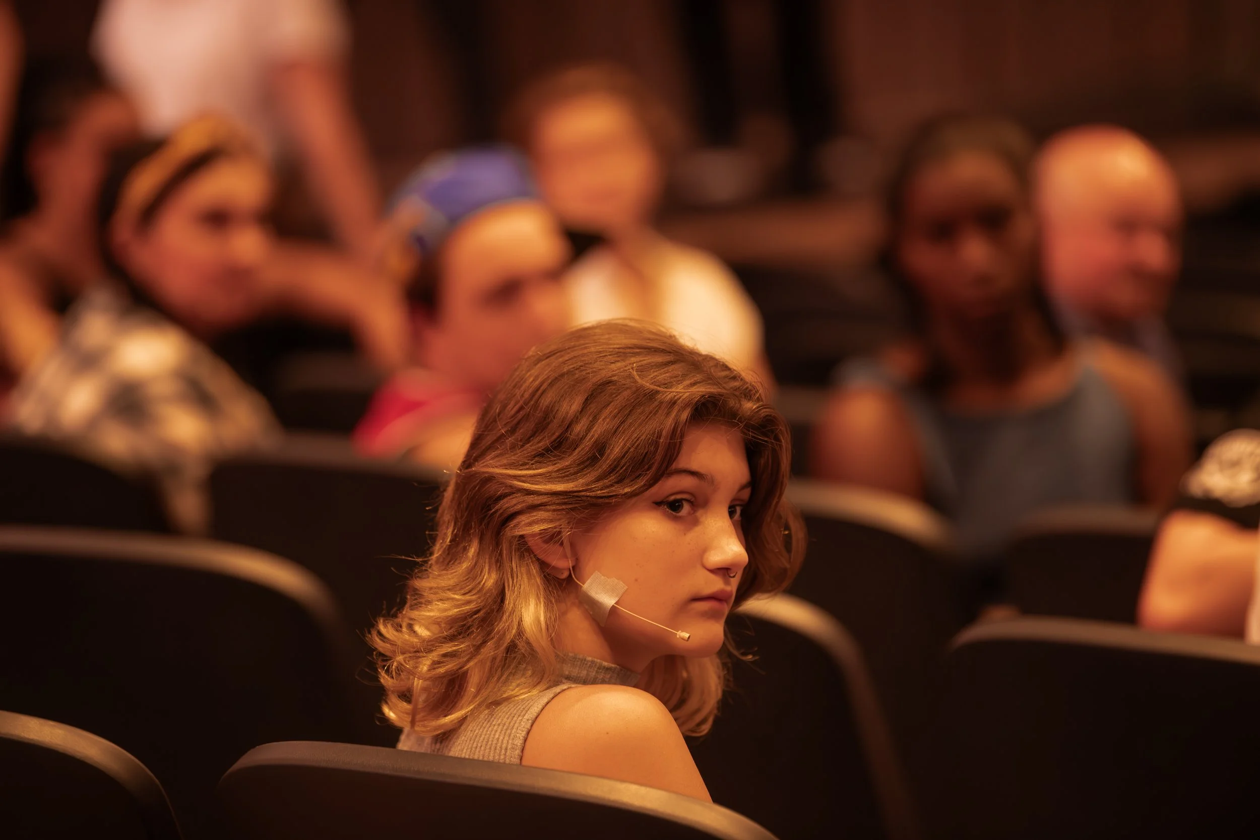 A young woman with shoulder-length wavy hair wearing a beige top and a headset, sitting in an auditorium among other attendees, looking over her shoulder.