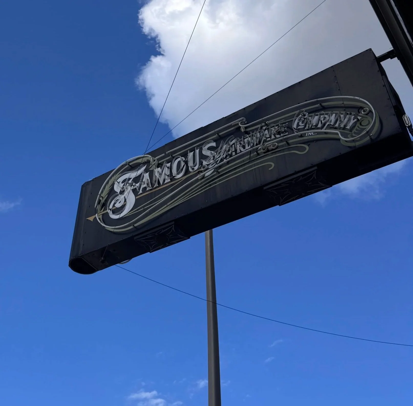 A black sign with the word 'FamousArnou' in stylized silver lettering, mounted on a pole against a bright blue sky with scattered white clouds.
