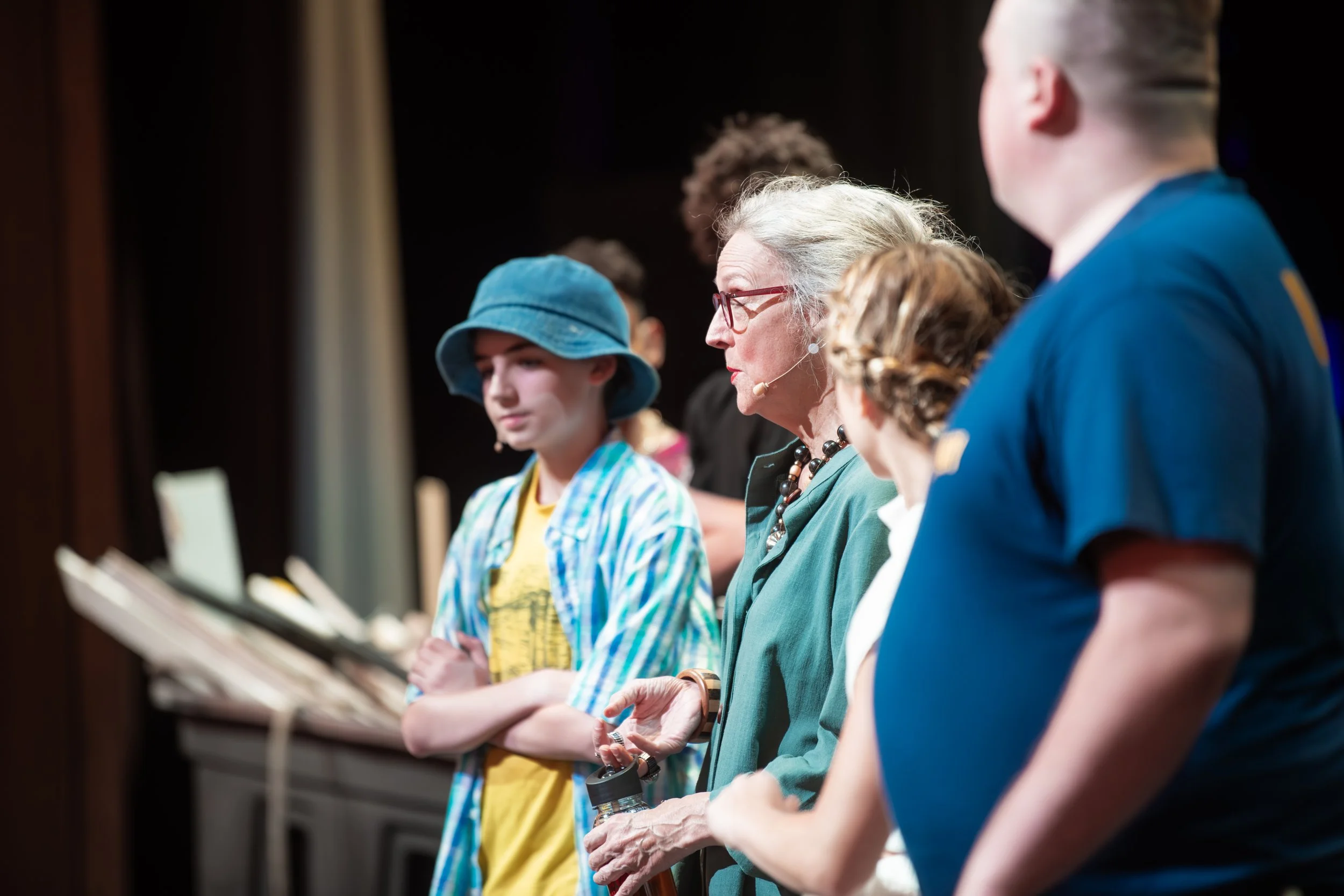 A diverse group of people, including a woman with glasses and a young girl with a blue hat, standing on a stage during a performance or presentation.