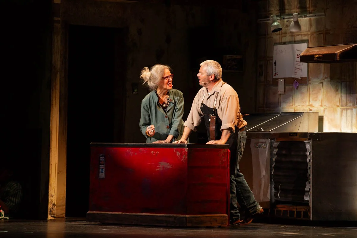Two actors performing on stage, engaged in a conversation, in a rustic kitchen set.