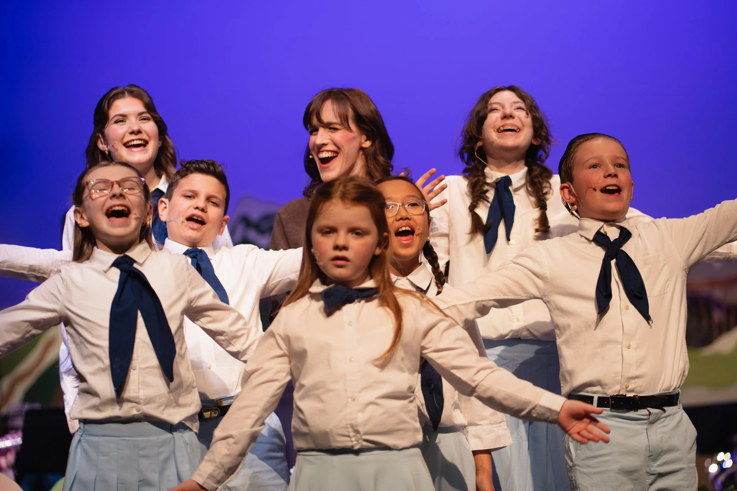Children and adults singing or performing on stage, smiling and raising arms in a scene from a musical or theatrical performance.