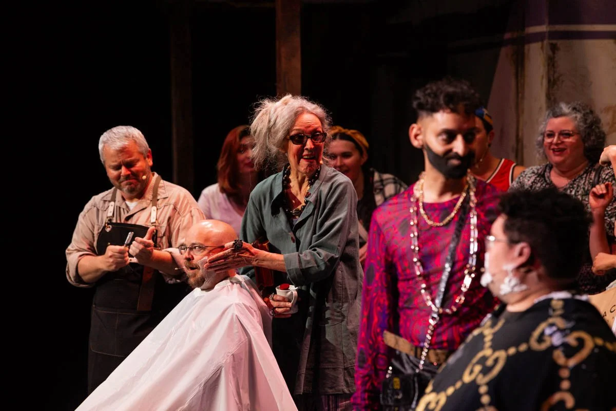People on stage during a theatrical performance, with a woman in glasses and gray hair doing makeup or costume work on a seated man with a bald head and beard, others watching and smiling in the background.