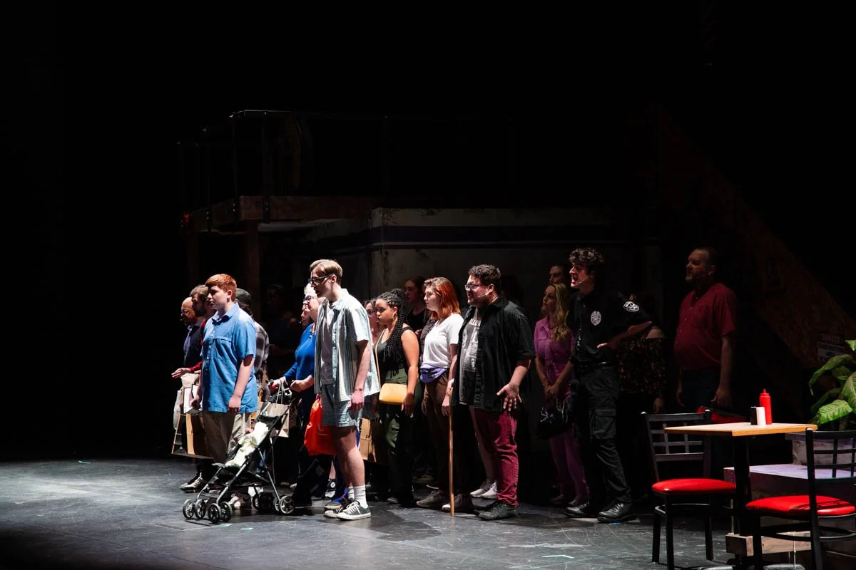 A group of diverse people standing in a line on a stage in a dark theater, with a spotlight illuminating them, and some tables and chairs nearby.