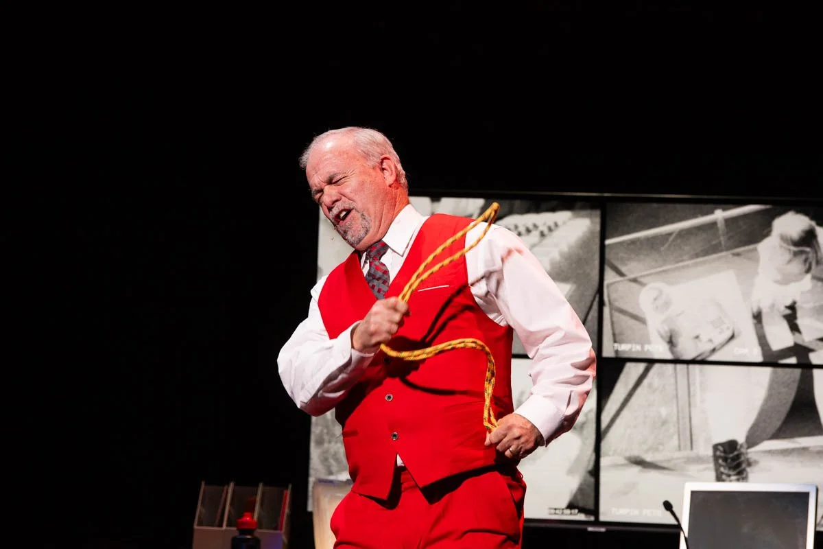 Man with gray hair and beard performing on stage, wearing a red vest over a white shirt, holding a yellow rope, with black and white images in the background.