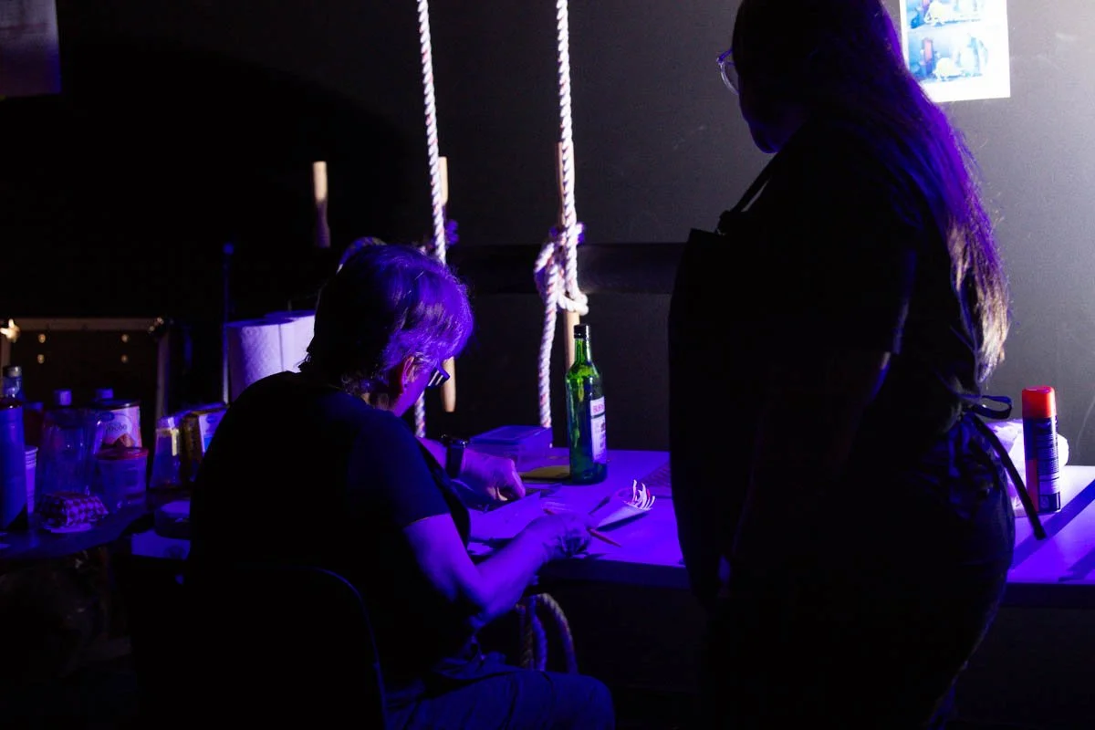 Two women in a dimly lit room with black walls, one sitting at a table with a green bottle and some papers, and the other standing nearby. The room has hanging ropes and various objects on the table, including bottles and a can.