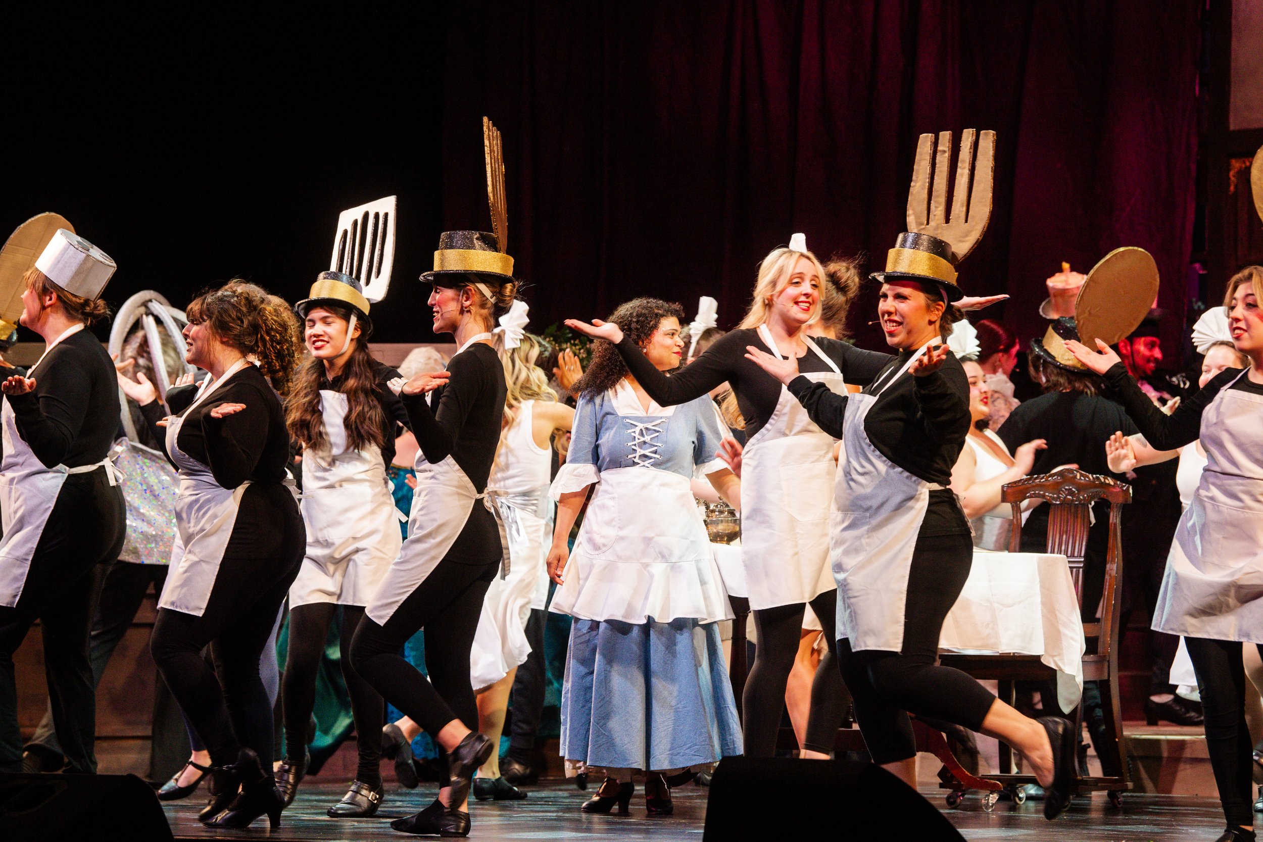 Stage scene with performers dressed as chefs, wearing hats and aprons, dancing in a line, some with large fork and spatula props on their heads, in a theater production.