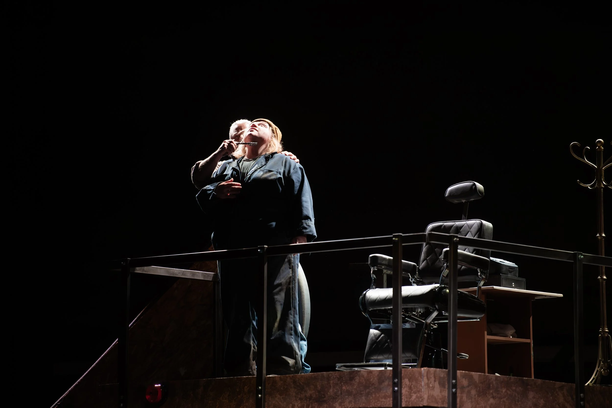 Two performers, one older man with a beard, and a woman, on a dark stage, illuminated by a spotlight, with empty office chair and furniture around them.