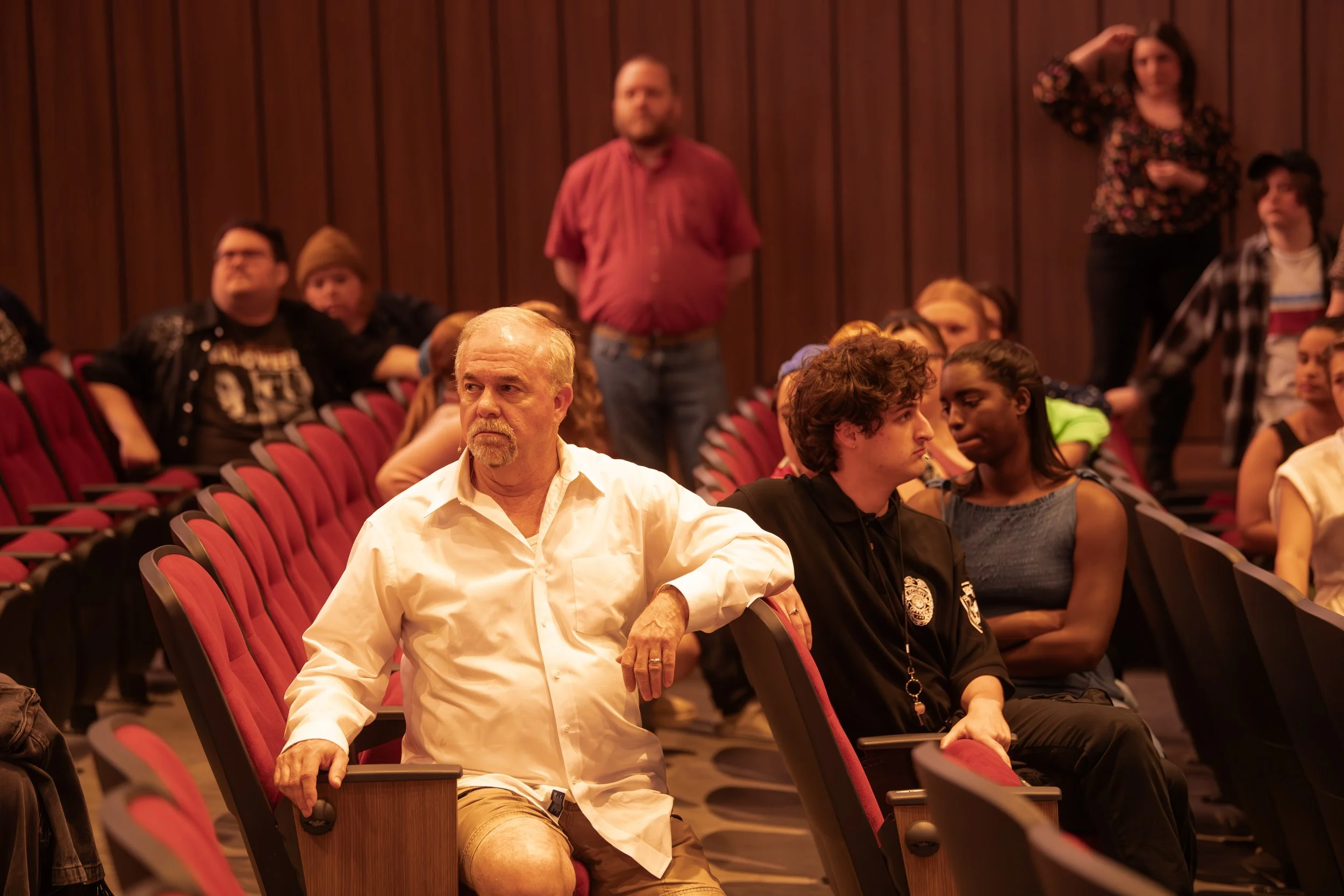 People sitting in an auditorium, listening attentively. The front row has an older man and a young man in uniform, with other audience members behind them.