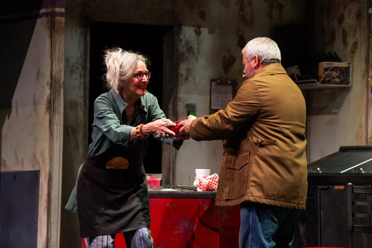 A woman with gray hair and glasses, wearing a gray shirt and a black apron, is smiling and handing something to a man with gray hair, wearing a brown jacket, in a rustic setting with worn walls and shelves.