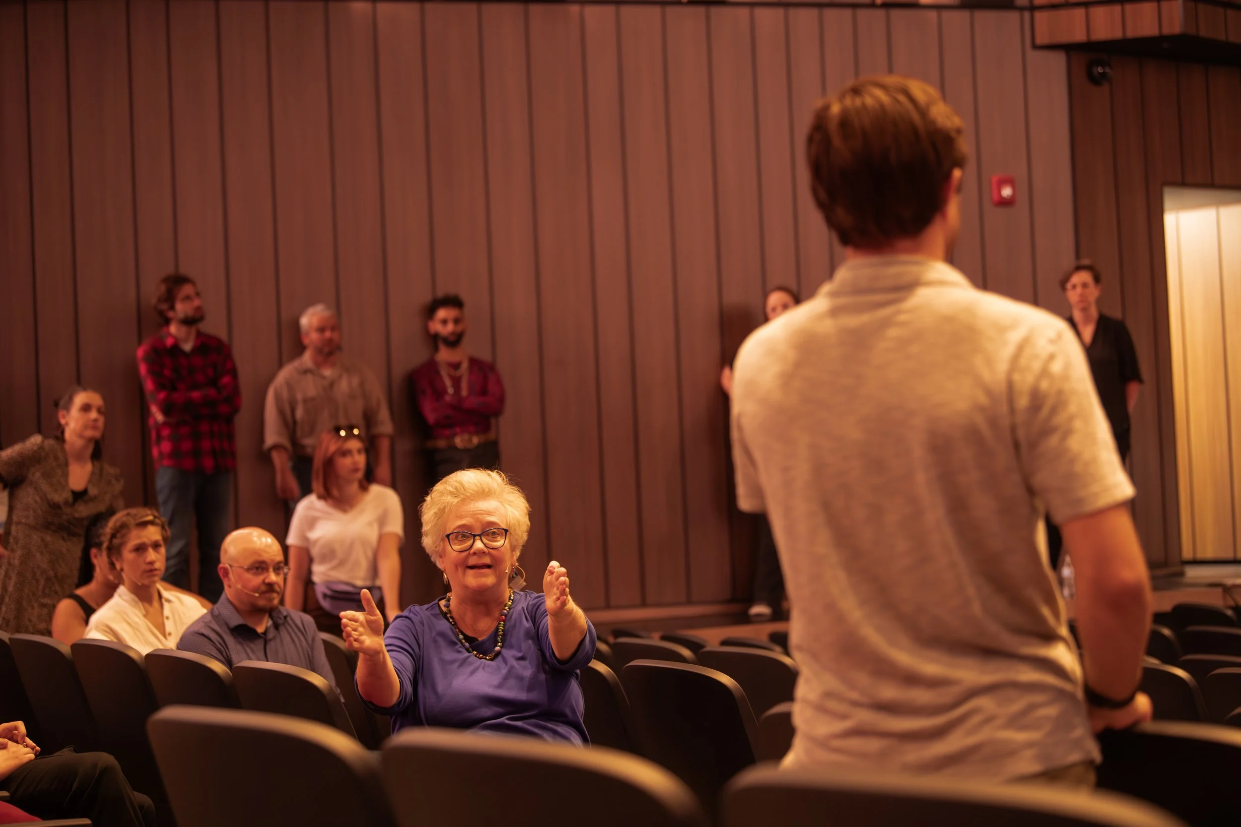 An elderly woman with glasses and a blue shirt is gesturing with her hands while talking to a young man in a light-colored shirt in a theater or auditorium. Several other people are seated and standing, listening to the interaction, with a wood-panel