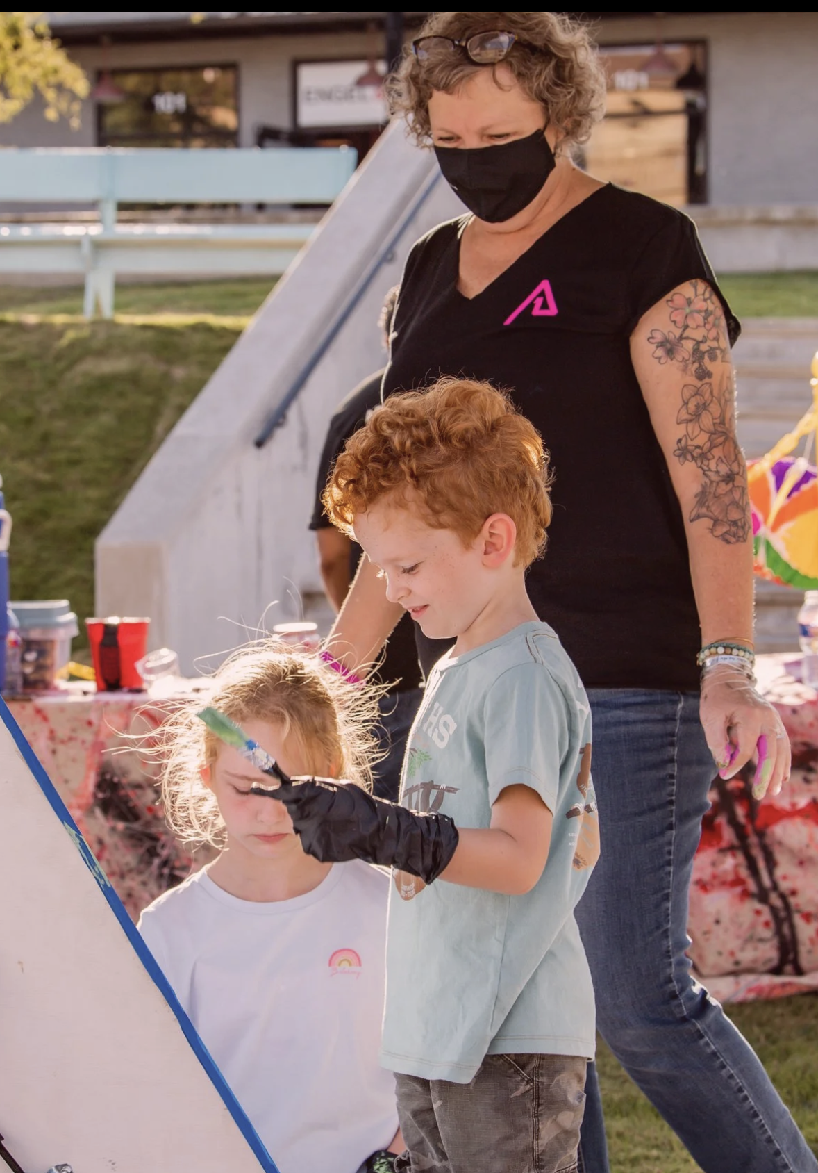 Adult woman with short, curly hair, wearing a black face mask and a black shirt with a pink logo, standing outdoors with two children, one boy and one girl. The woman has floral tattoos on her arm and is holding pink and purple objects. The children have curly red hair and light skin, and the girl is being painted or marked on her face with a black glove on her hand. The scene appears to be at an outdoor event or fair.