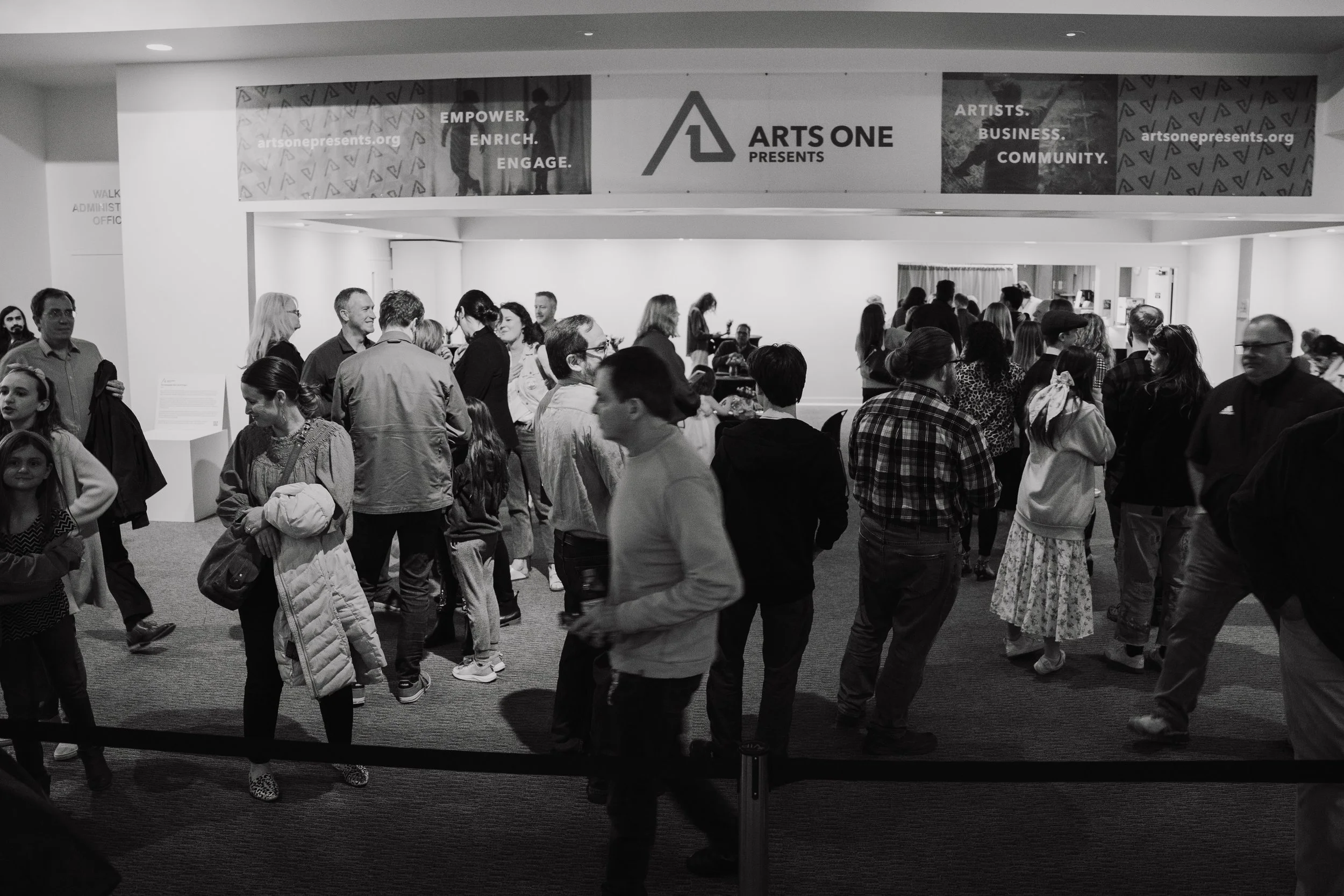 People gathering at an art event in a lobby, with a large banner reading 'Arts One Presents' and slogans like 'Empower, Enrich, Engage' displayed overhead.