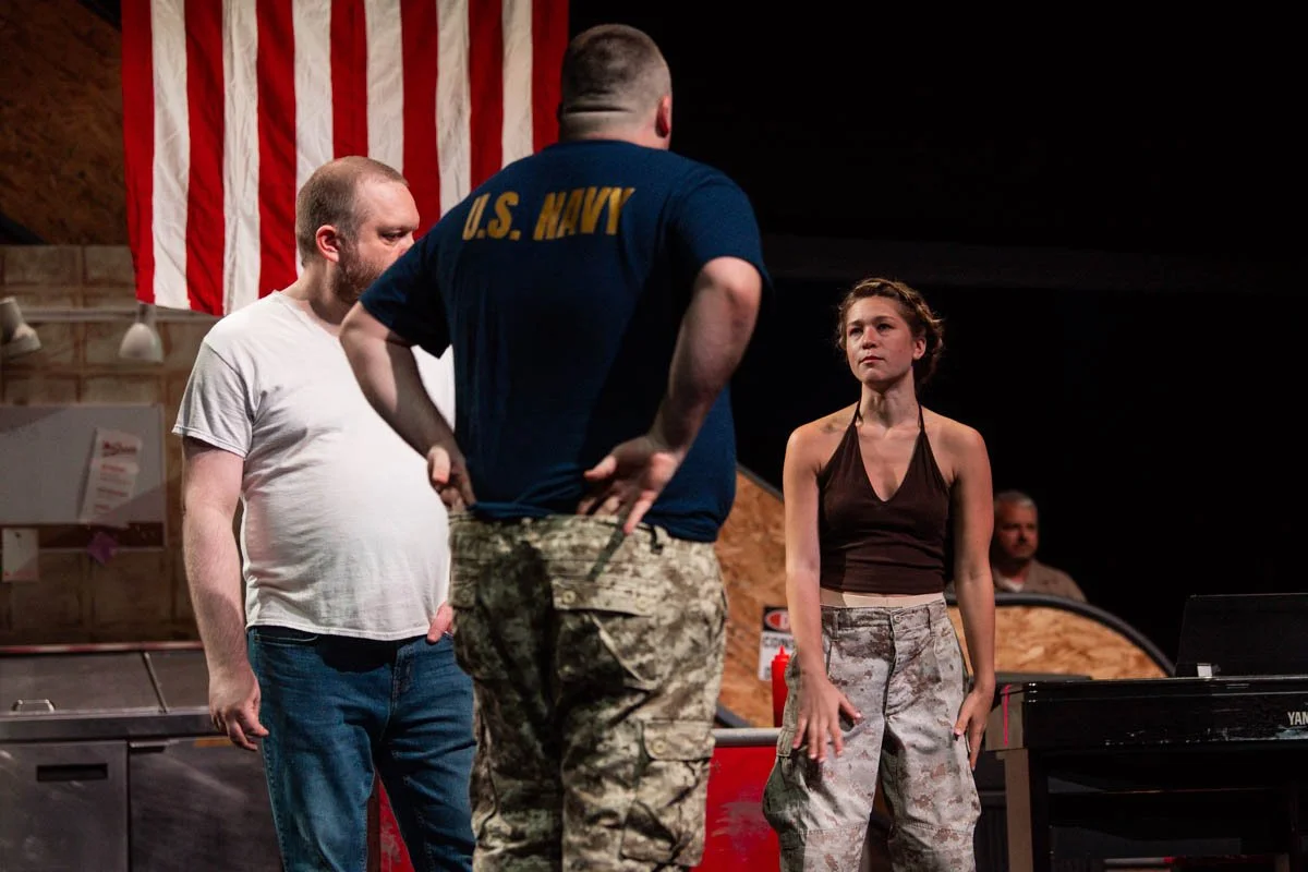 Three people standing indoors, with one person facing away, wearing a U.S. Navy shirt, and two others facing forward, one in a white t-shirt, and the woman in a dark halter top, with an American flag hanging in the background.