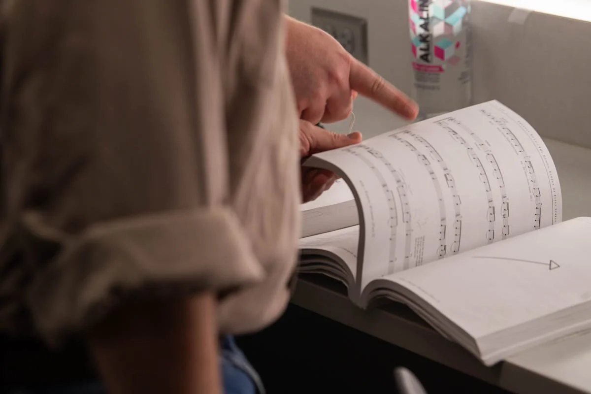 Person flipping through sheet music or a music book on an airplane tray table, with a can of ALO drink nearby.