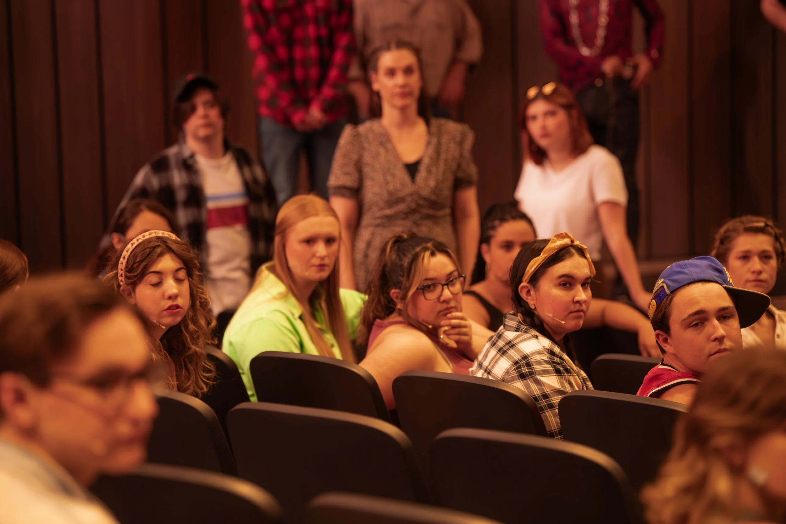 A group of young people seated in an auditorium or theater, with some looking forward and others appearing bored or distracted, and a woman standing at the back.